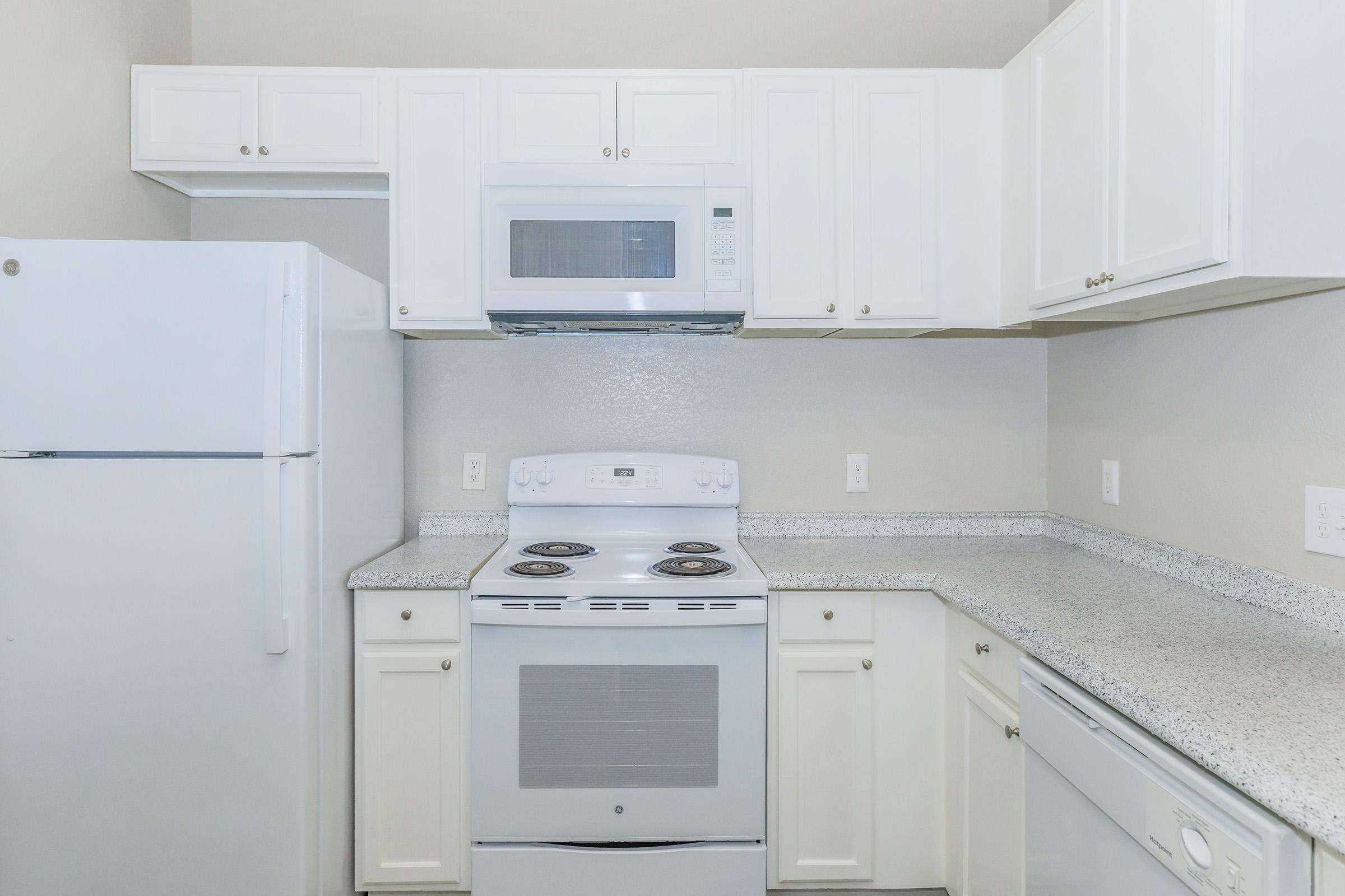 A modern kitchen featuring white cabinets, a white refrigerator, a built-in microwave above the stove, and a smooth-top electric range. The countertops are light-colored with a speckled design, and the walls are a neutral shade, creating a clean and contemporary look.