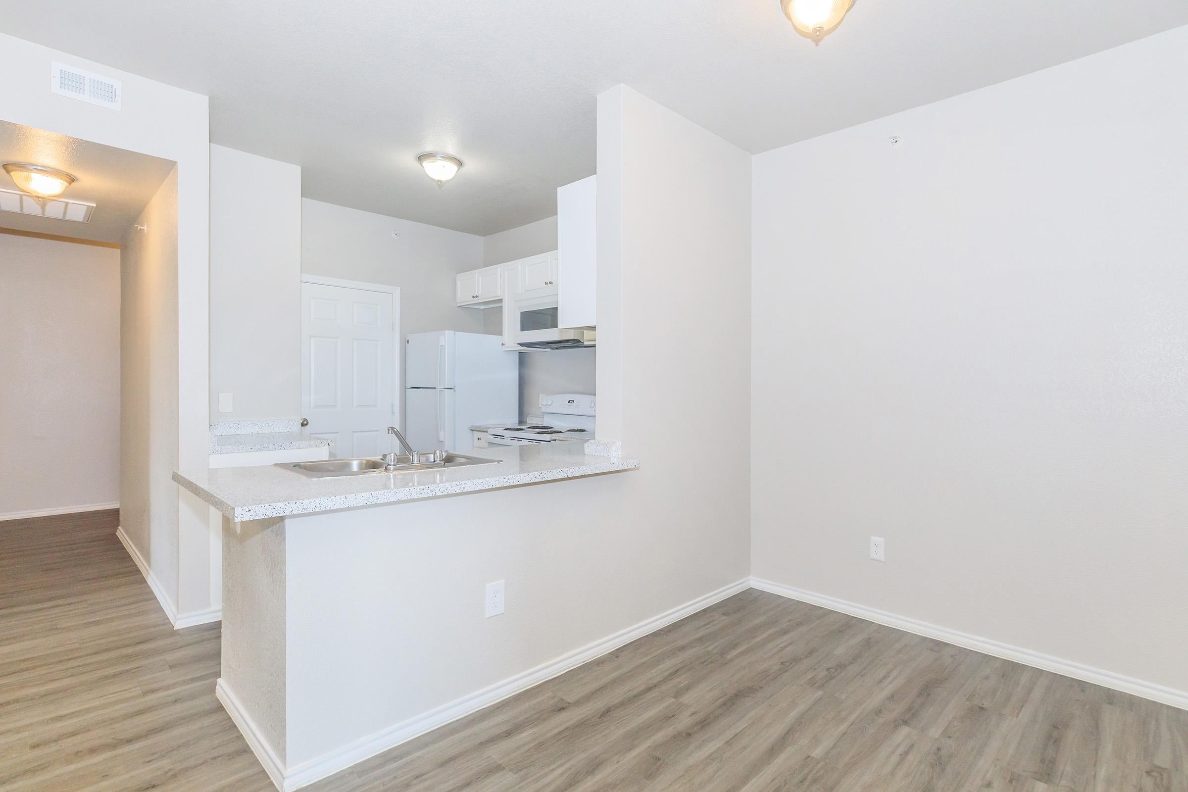 A modern kitchen and living area featuring light-colored walls and wooden flooring. The kitchen includes white cabinetry, a stove, and a refrigerator. A countertop with a sink separates the kitchen from the living space, creating an open layout. Two ceiling lights provide illumination.