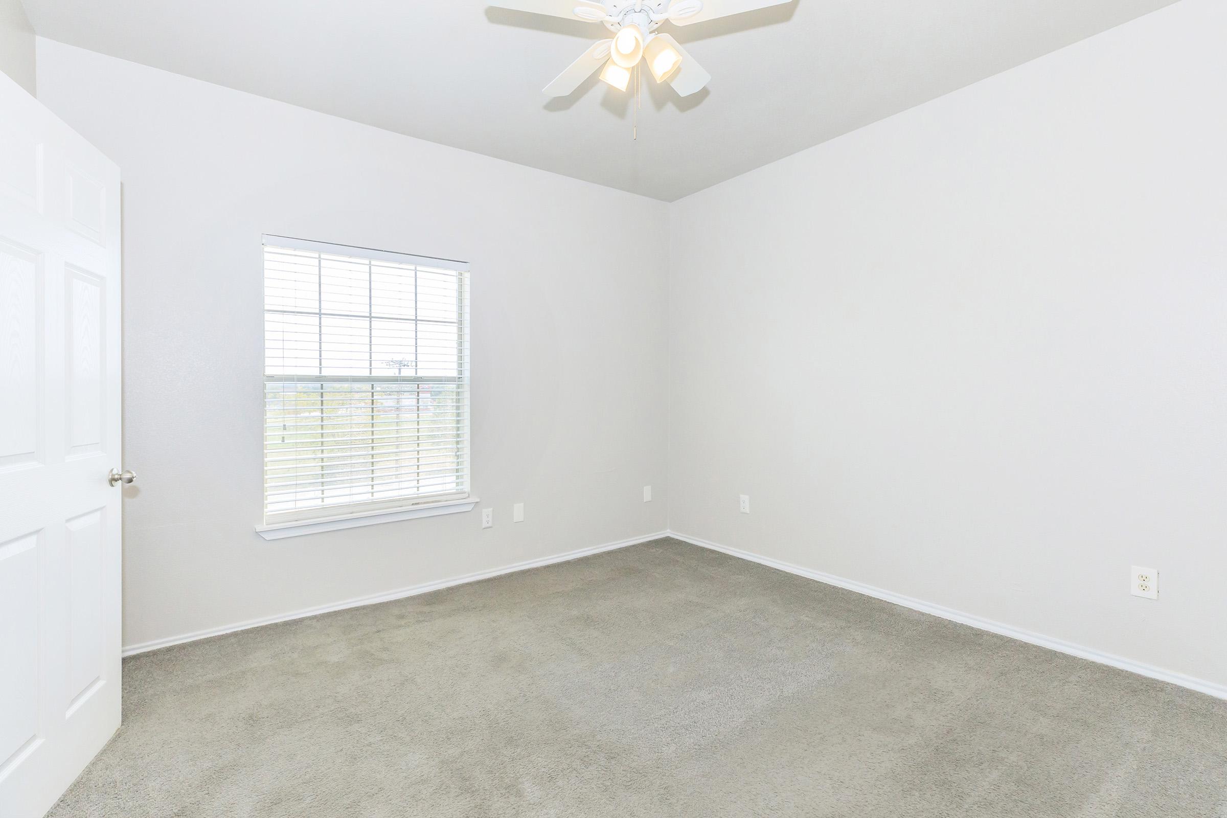 Empty room with light gray walls, a ceiling fan, and a window with white blinds. The floor is covered with light gray carpet. There is a white door on the left side of the image, and the space is brightly lit, giving a clean and open feel.