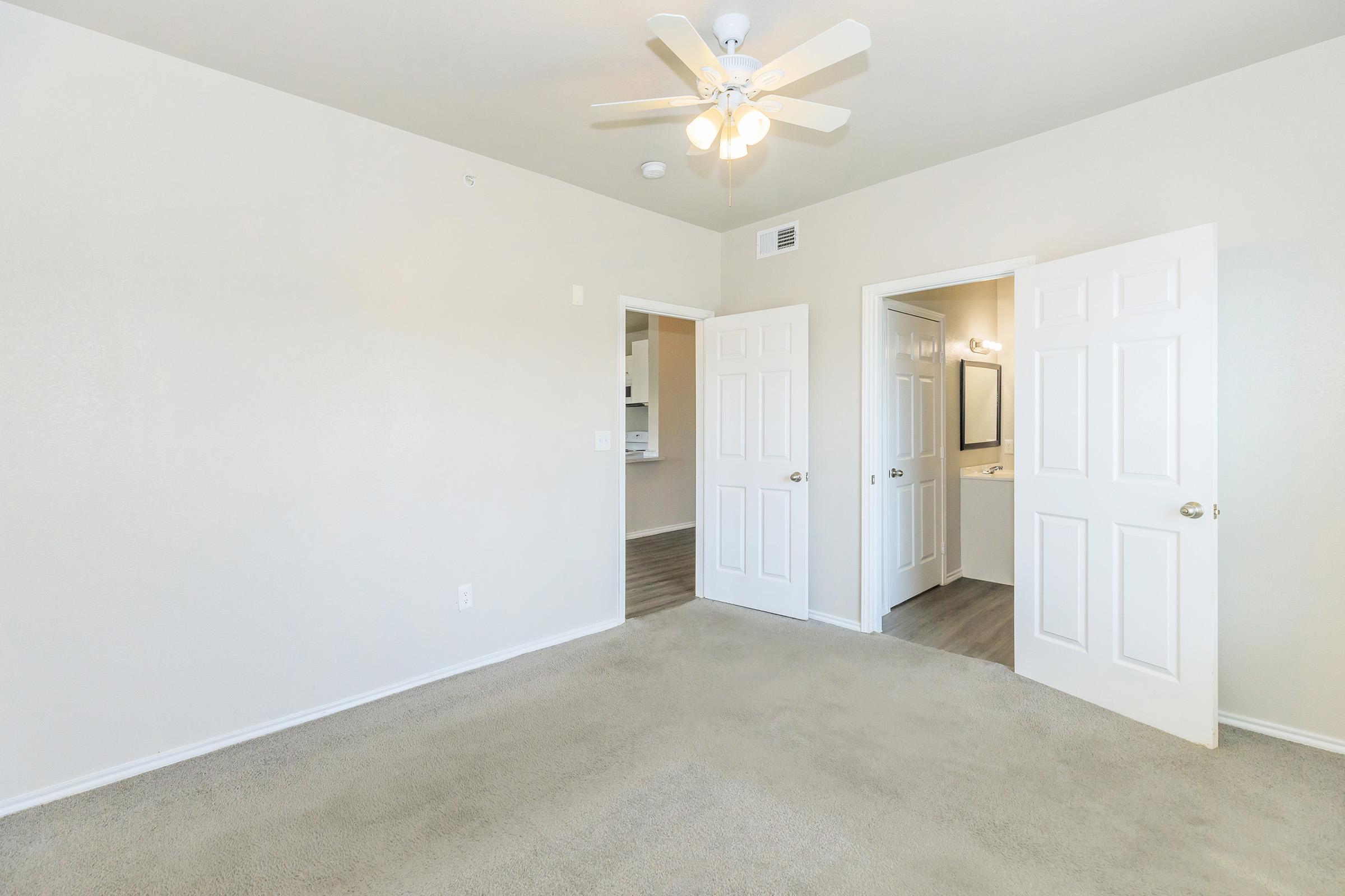 A spacious and well-lit room featuring beige carpet and light-colored walls. There is a ceiling fan, and two doorways lead to adjacent areas, one visible bathroom space and another room. Natural light flows in, creating a welcoming atmosphere.