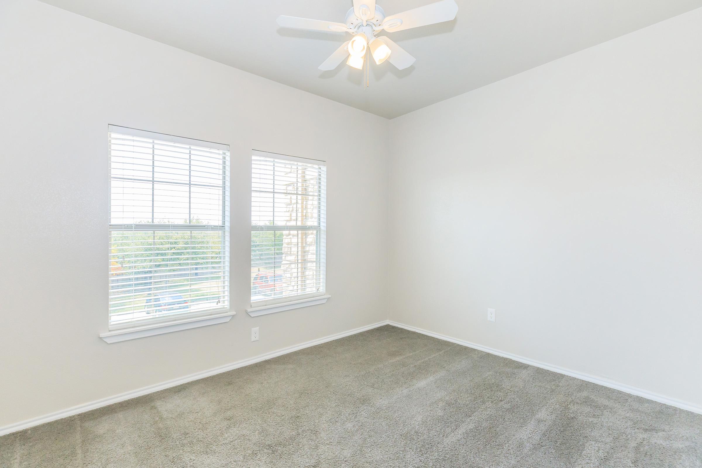 Empty room featuring gray carpet, two windows with white blinds allowing natural light, and a ceiling fan. The walls are painted a light color, contributing to a bright and open atmosphere. No furniture or decorations present.