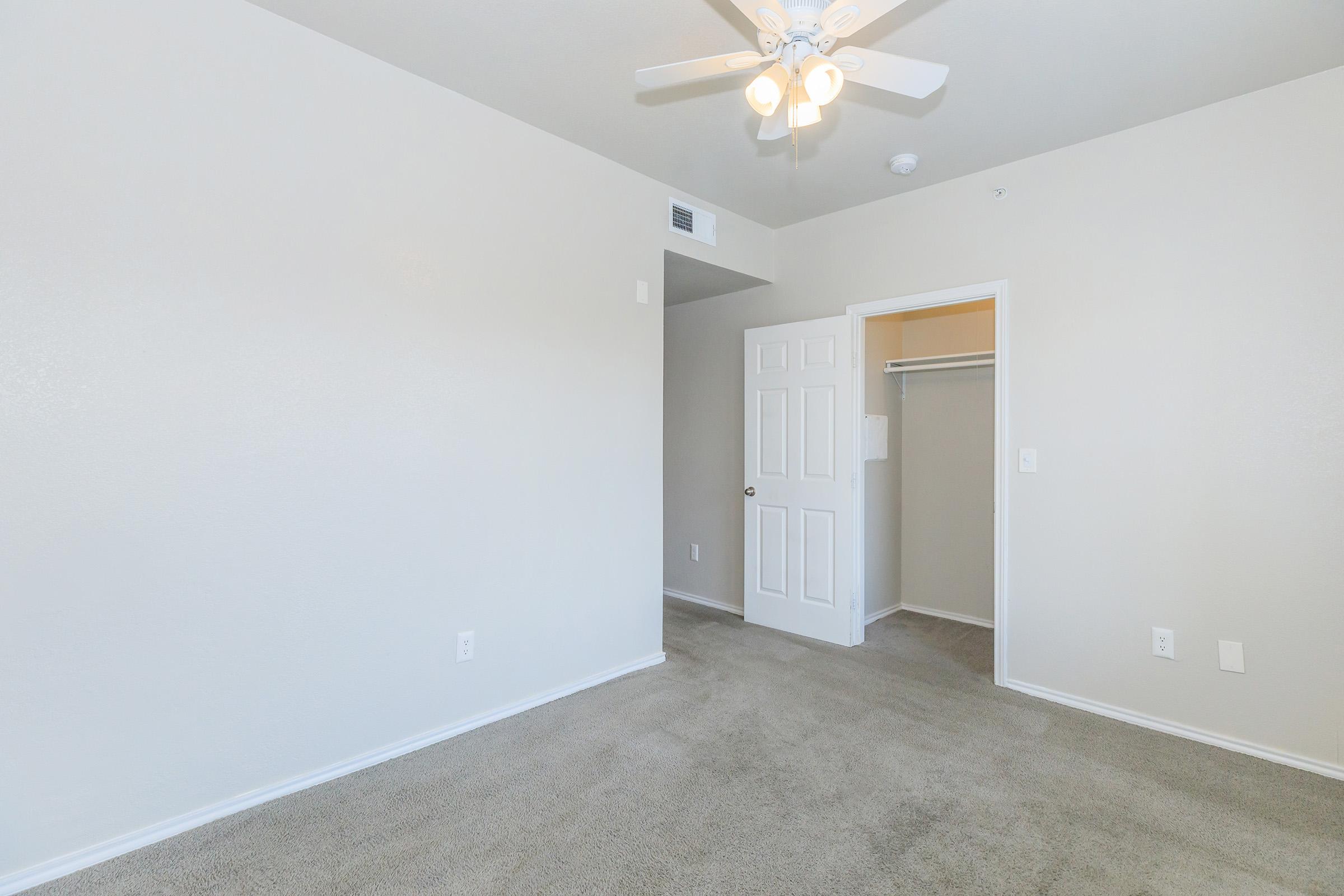 A spacious, empty bedroom with light beige walls and a ceiling fan. The floor is carpeted in a light color. A closed white door leads to a small closet, and there are no furnishings or decor, creating a minimalist and airy atmosphere.