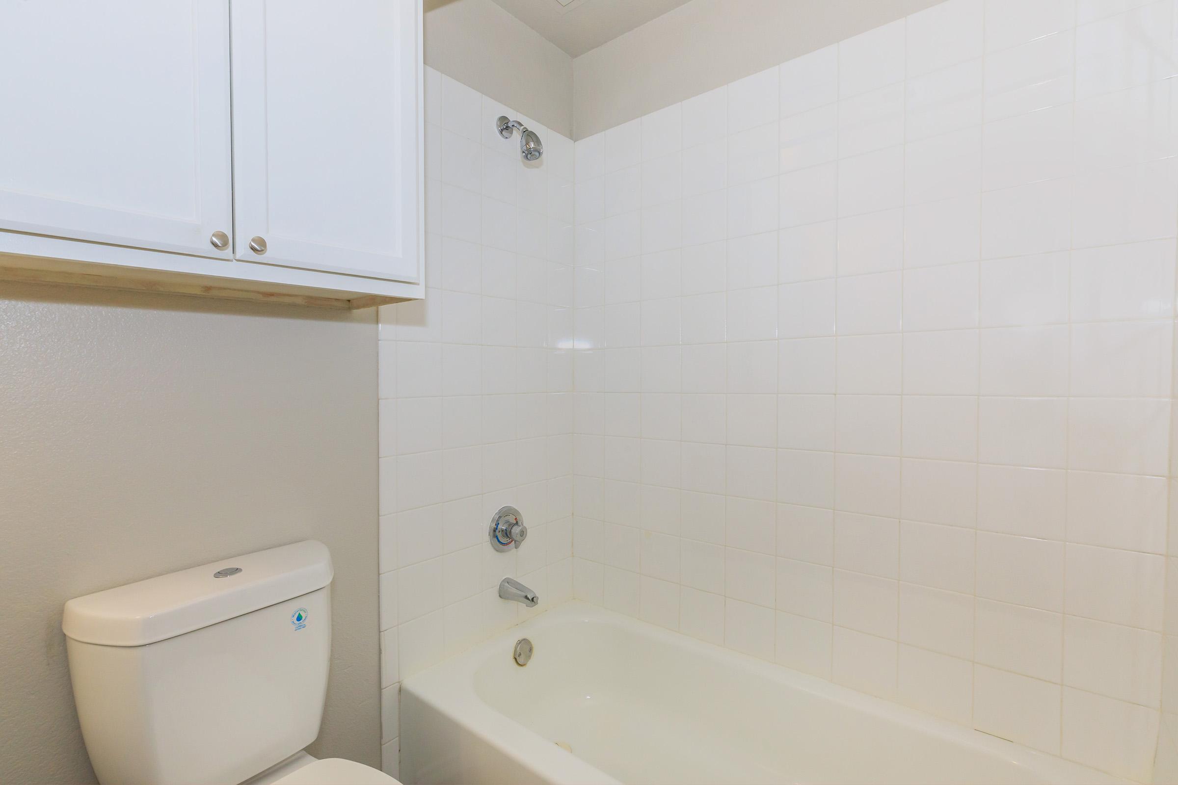 A clean bathroom featuring a white bathtub, a wall-mounted showerhead, and a white toilet. Above the tub, there are white tiled walls and a wooden shelf. The overall color scheme is neutral with light grey and white tones, creating a simple and modern look.