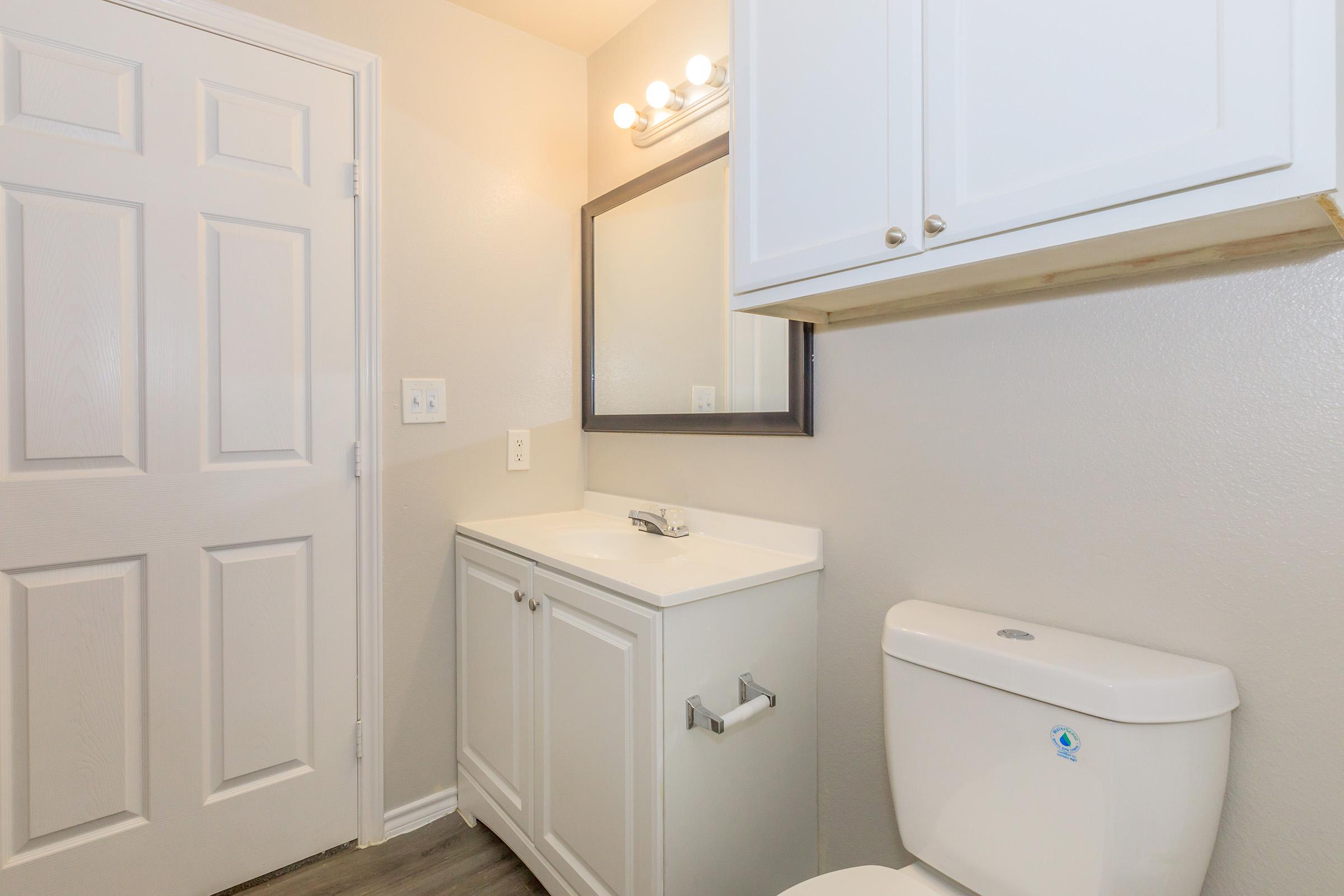 A clean and modern bathroom featuring a white vanity with a sink and a large mirror above it. To the right, there is a toilet. The walls are painted in a light color, and there is a door leading out of the room. The flooring is a light-colored wood laminate.