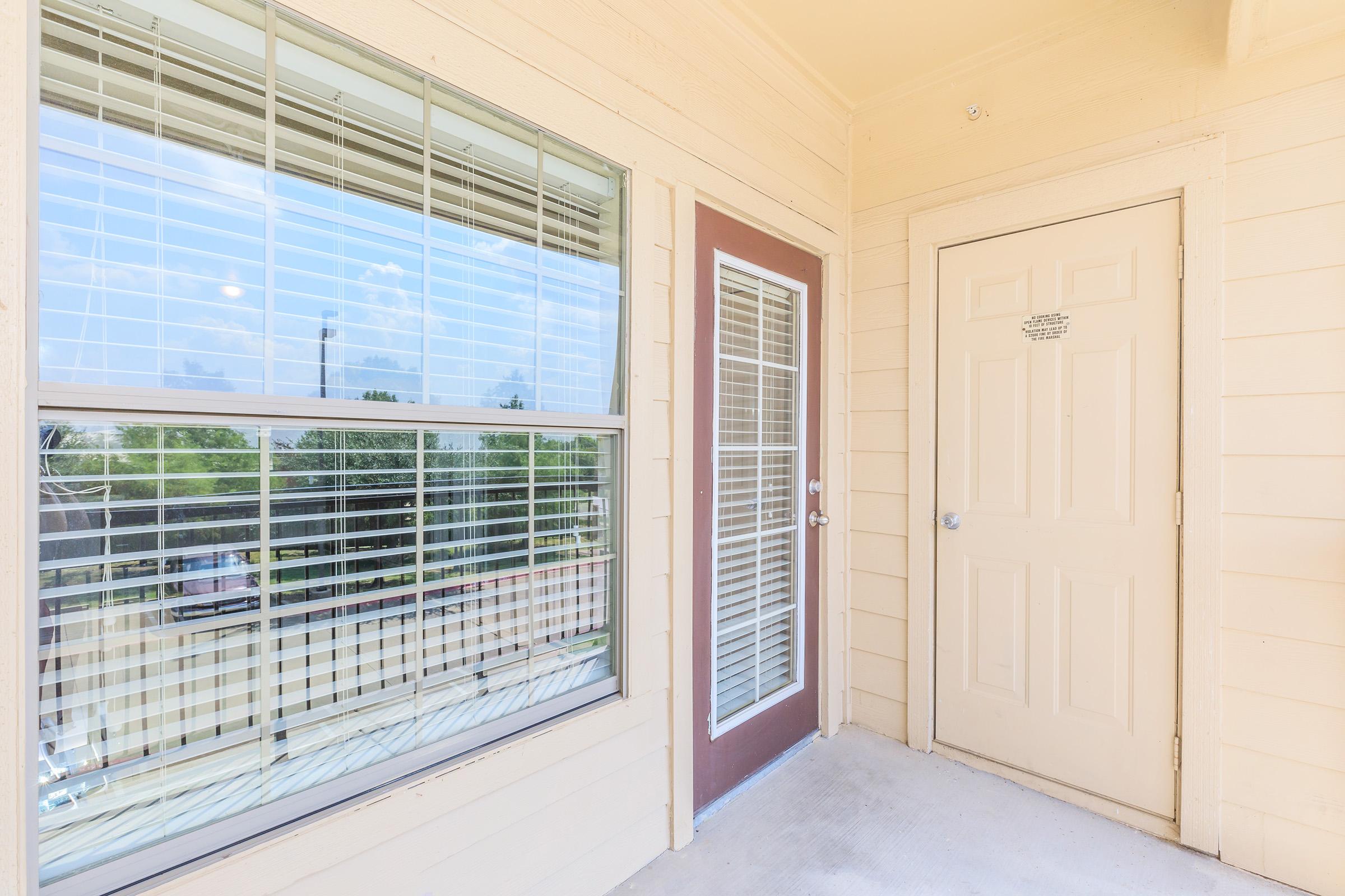 Exterior view of a light-colored apartment entryway featuring a door and a large window with horizontal blinds. The door is beige with a simple handle, while the window offers a view of greenery outside. The area is well-lit, showcasing a clean and welcoming atmosphere.