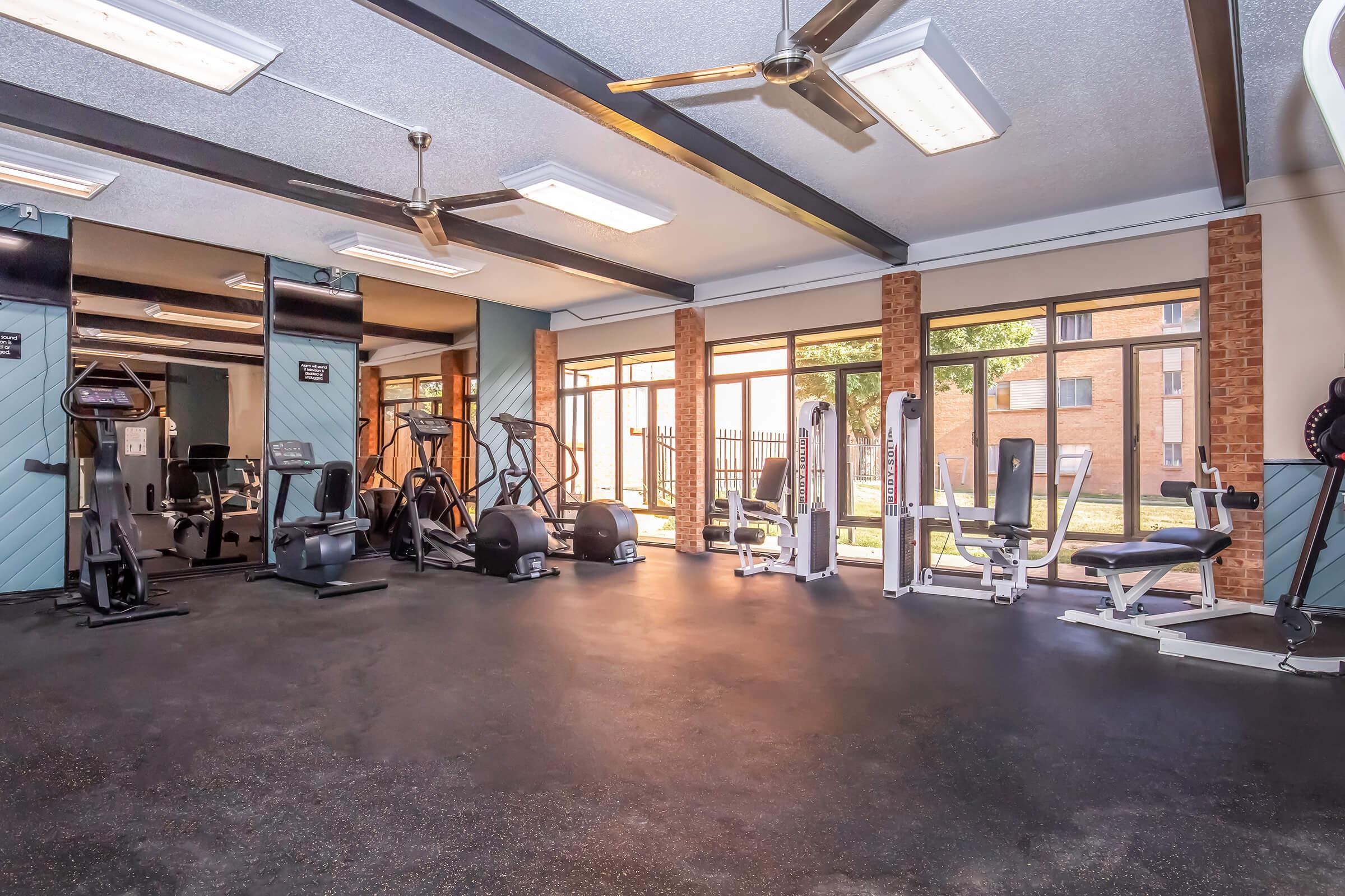 Interior of a fitness center featuring various exercise equipment, including cardio machines and weight machines. Large windows allow natural light to fill the space, creating a bright and inviting atmosphere. Fans are installed on the ceiling for ventilation.