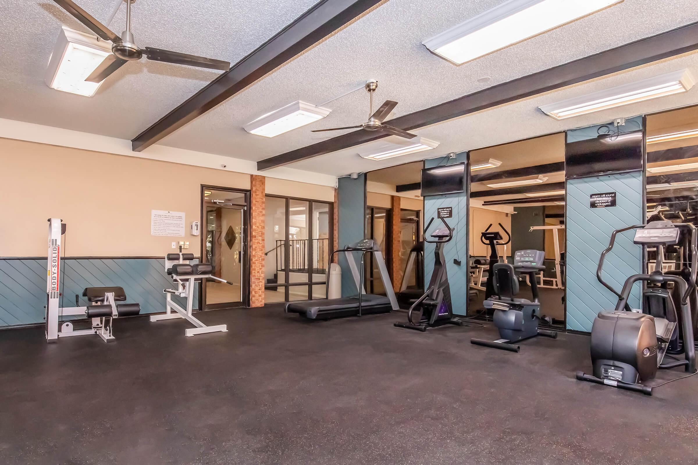 A well-equipped gym featuring various exercise machines, including weight benches, treadmills, and a stationary bike. The space has a polished floor, ceiling fans, and large mirrors on the walls. Natural light streams in through the windows, creating an inviting workout environment.