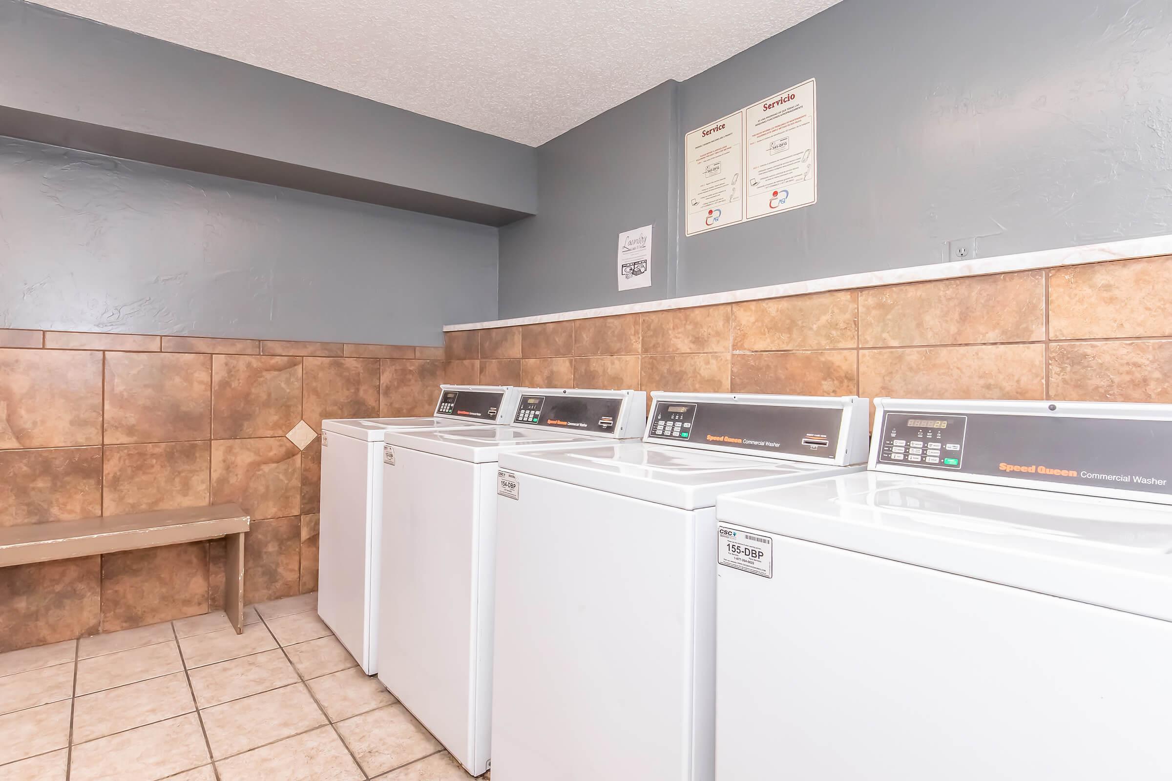 A laundry room featuring four commercial washing machines lined up against a wall. The area has beige tiled flooring and walls painted in a gray color. There is a bench on the left side, and informational signs are posted on the wall.