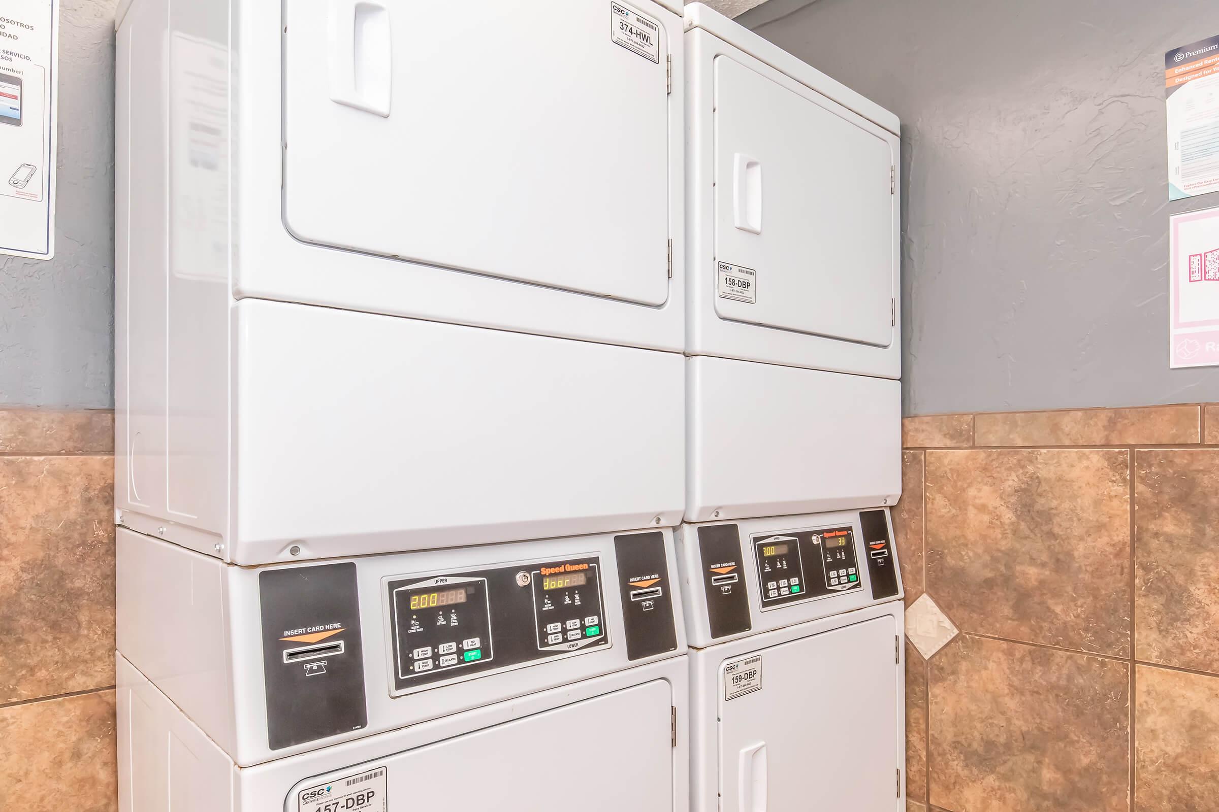A stack of white coin-operated dryers with control panels on the front, located against a textured wall. The dryers are placed next to tiled flooring and there are posters or notices on the wall above.