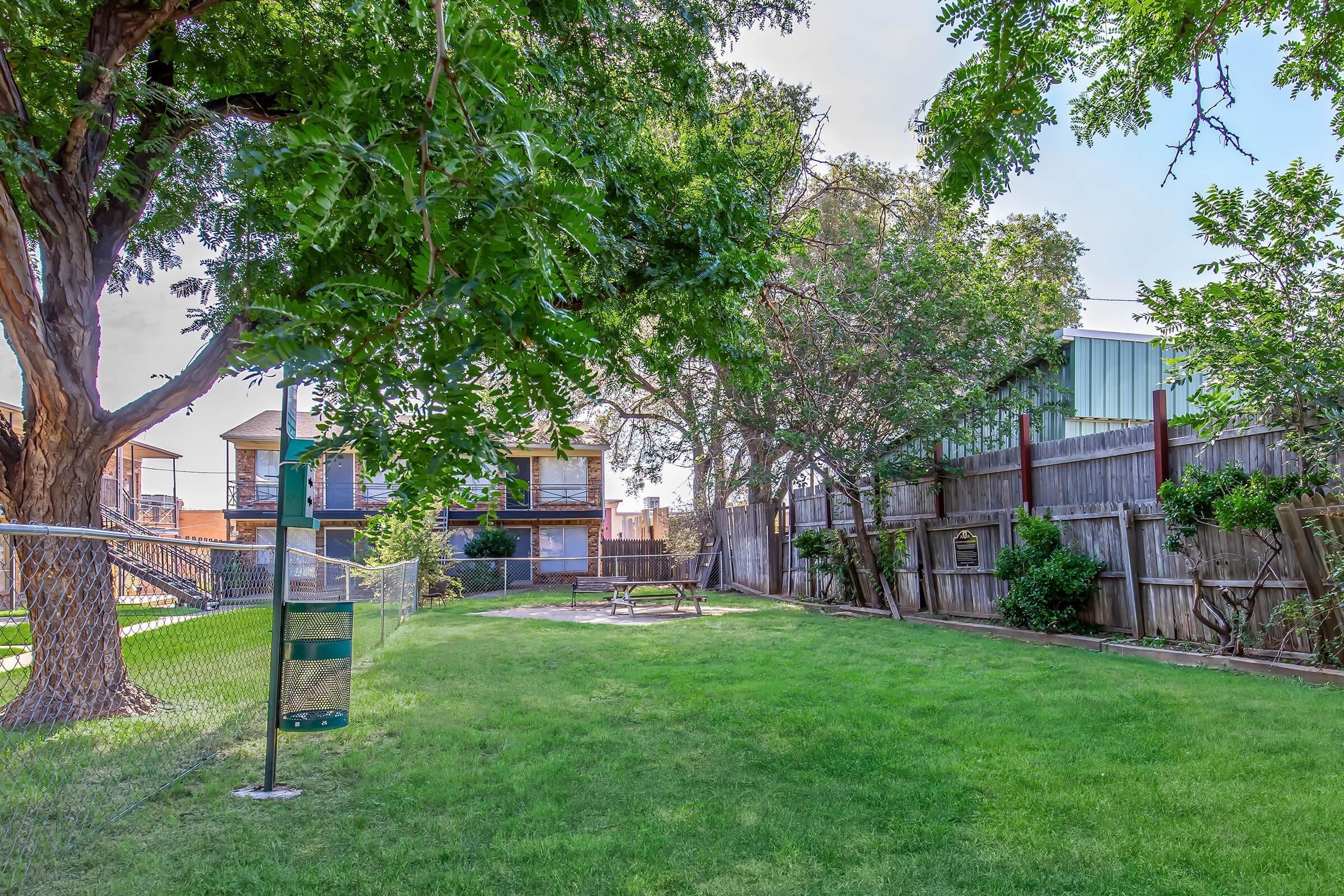 A green park area with a well-maintained lawn, surrounded by trees. There are wooden benches, a playground area, and buildings in the background. A chain-link fence encloses the space, providing a relaxing outdoor setting.