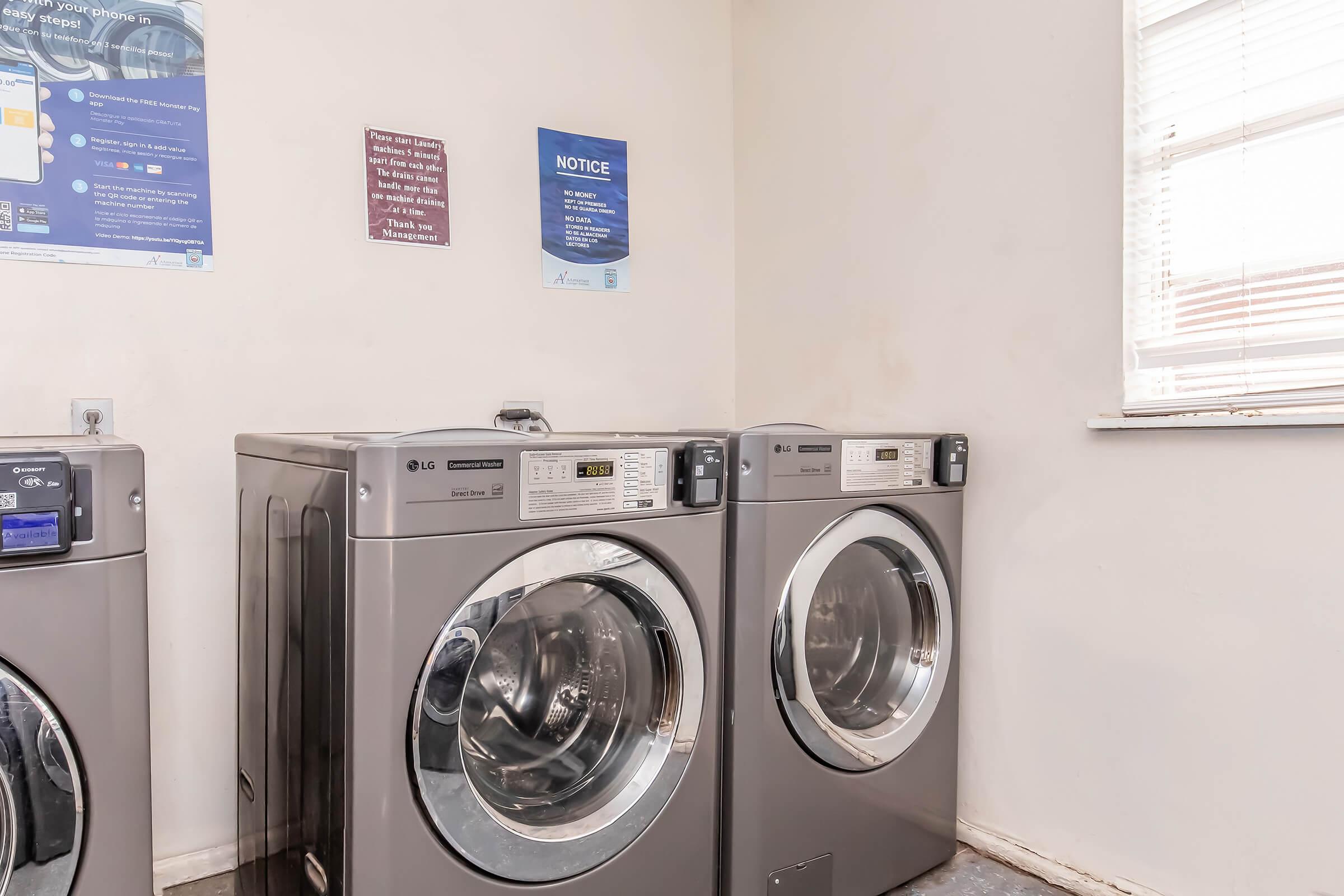Two gray front-loading washing machines side by side in a bright laundry room. The wall features notices and instructions above the machines, with a window providing natural light. The floor is bare, and the overall atmosphere is clean and functional.