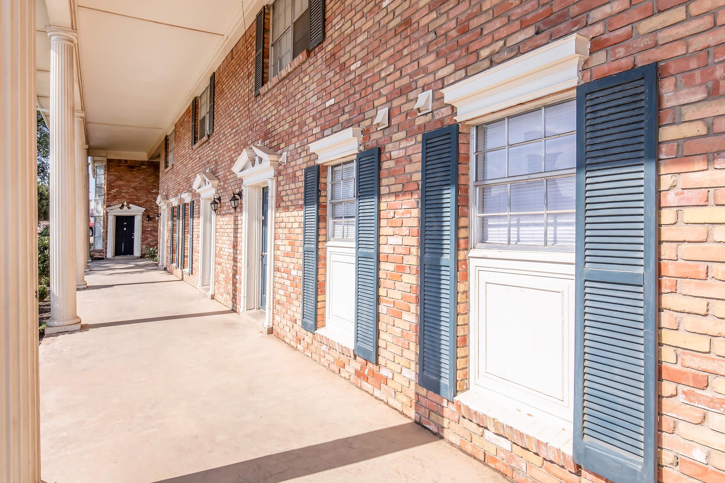 A view of a brick building's exterior, featuring several windows with blue shutters. The architecture includes classic columns and a covered walkway. The area appears well-maintained, with a clear path and some greenery visible in the background.