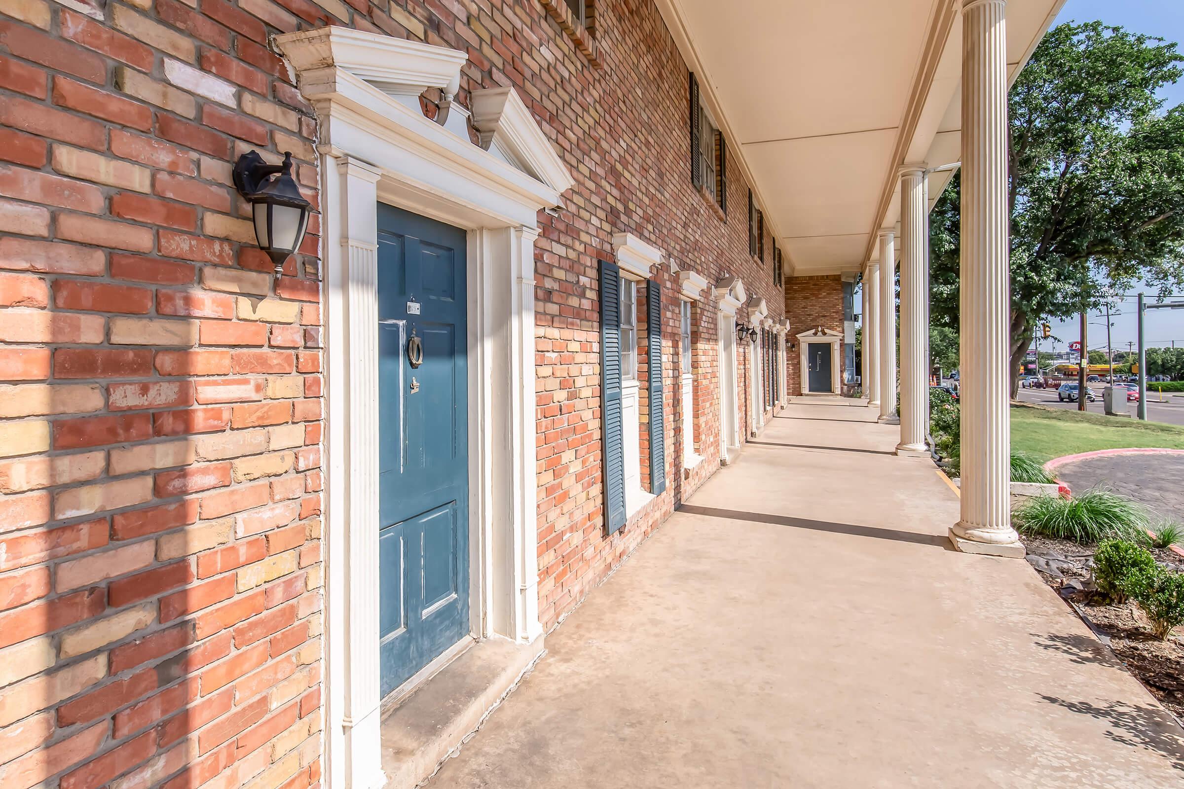 A brick building with a series of blue doors, lined along a covered walkway. The porch features tall white columns, and the exterior is well-lit by wall-mounted lights. The ground is a smooth, light-colored surface, and there are trees and greenery visible along the edges of the walkway.