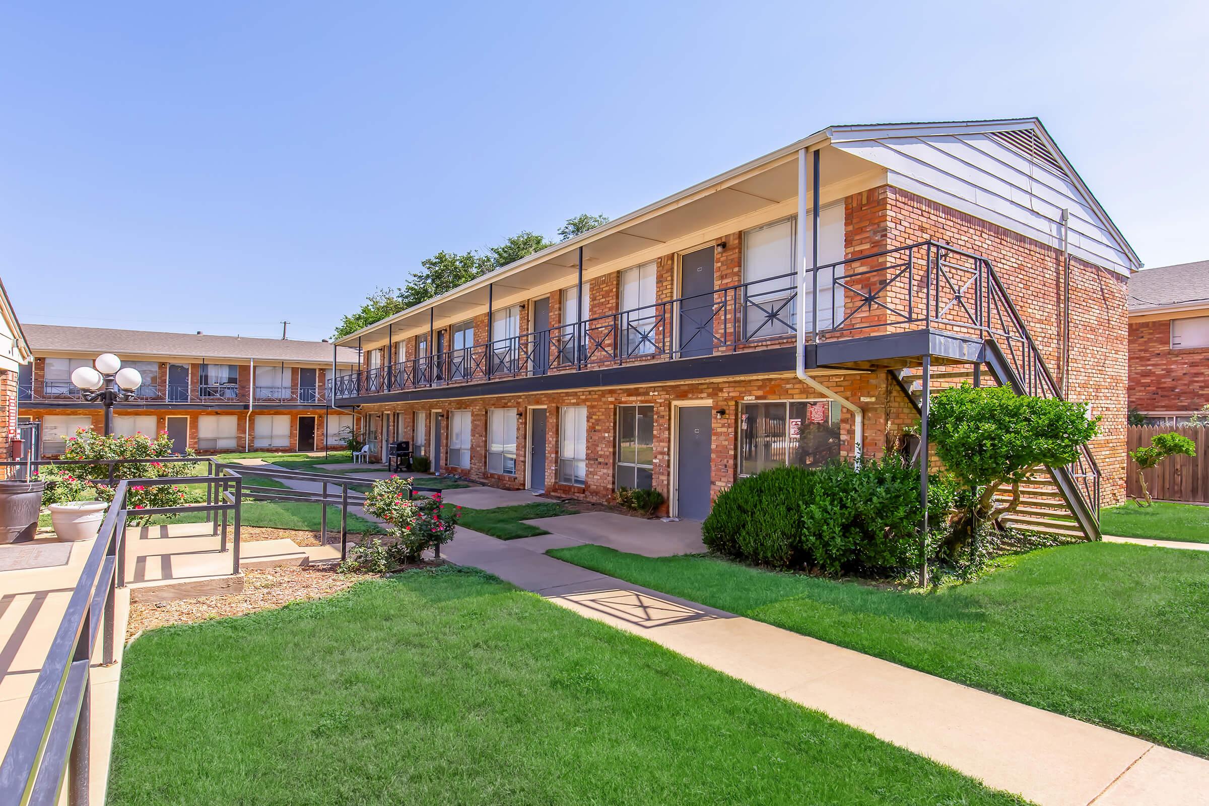 Two-story brick apartment building with multiple units. Each unit has a balcony and front door facing a landscaped courtyard. There are walkways and green grass areas, along with small shrubs and plants. Clear blue sky visible above.