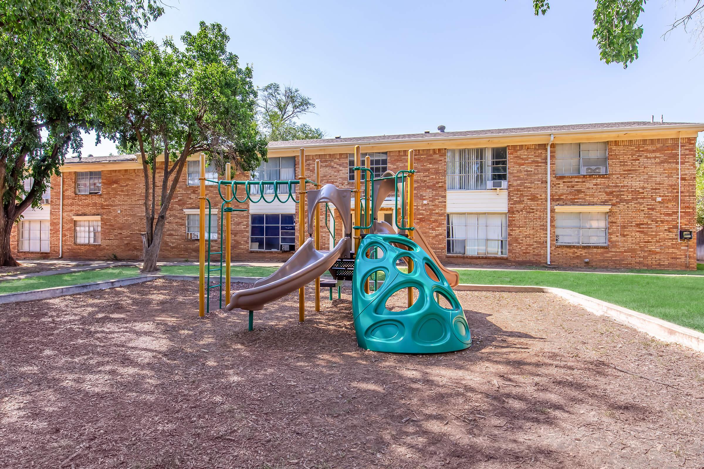 A playground featuring a colorful climbing structure with slides, surrounded by trees and grass. The background includes a brick apartment building. The area is well-maintained, providing a space for children to play.