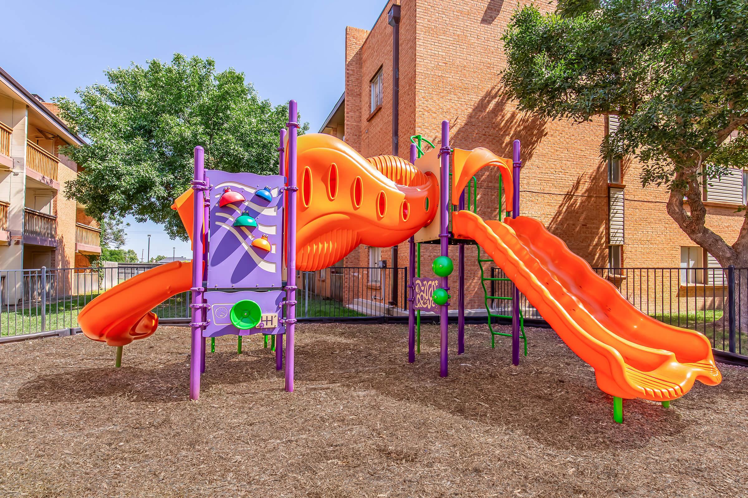 Colorful children's playground featuring two orange slides, climbing structures, and playful designs, set in a grassy area surrounded by multi-story brick buildings. The playground is enclosed with a fence, providing a safe play environment.