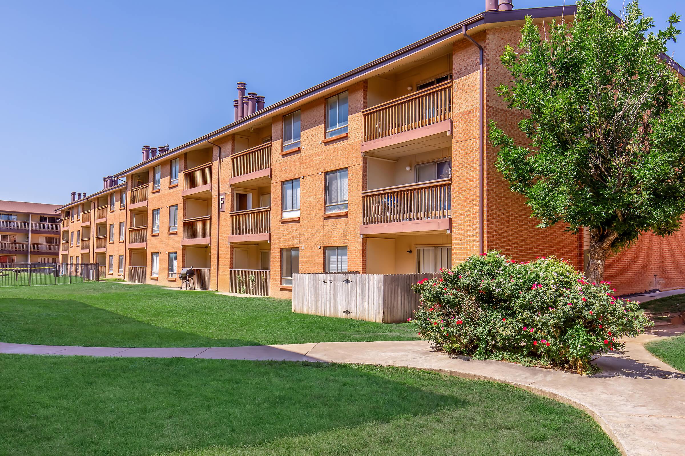 A view of a multi-story brick apartment building with balconies, surrounded by a well-maintained grassy area. In the foreground, a curved pathway leads to the building, and lush greenery including a bush with pink flowers is visible nearby. The sky is clear and blue.