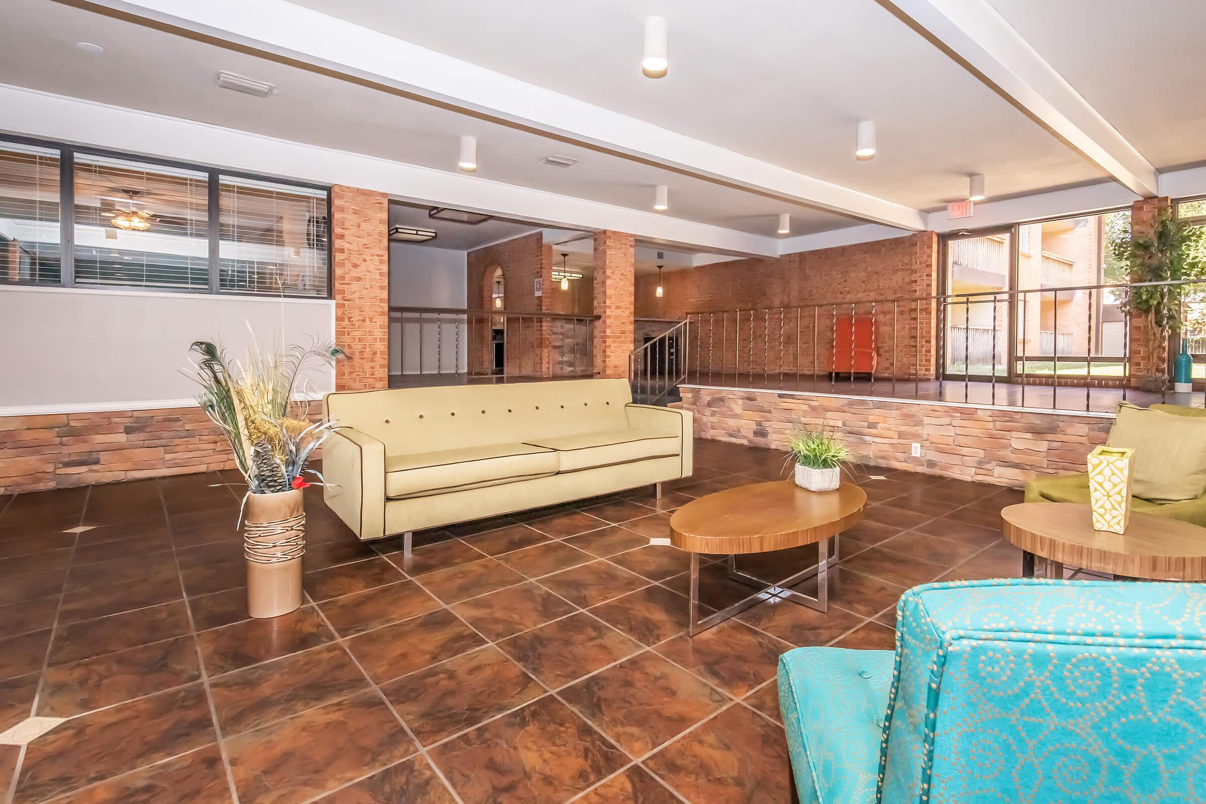 A modern lounge area featuring a beige sofa and round wooden coffee table. There are potted plants and decorative accents. The space has brick walls and large windows, providing natural light. The tile floor adds a contemporary touch, and there are railings in the background, suggesting multi-level architecture.