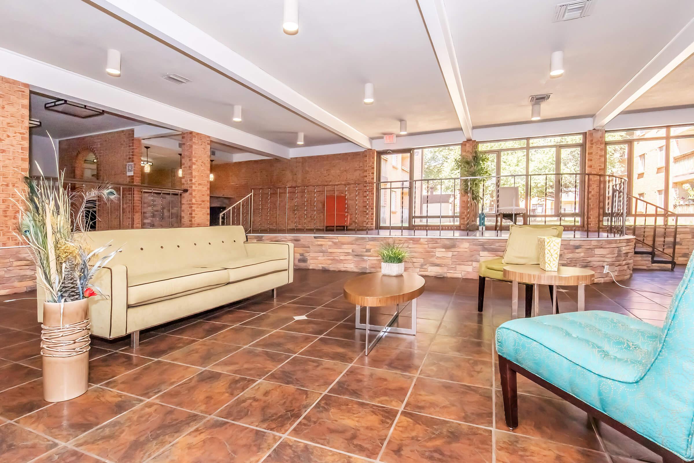 A cozy lobby area featuring a light green sofa, two round wooden tables, and a blue upholstered chair. The space has brick walls, tiled flooring, and large windows allowing natural light. Decorative plants and a small lantern add warmth to the modern, inviting atmosphere.