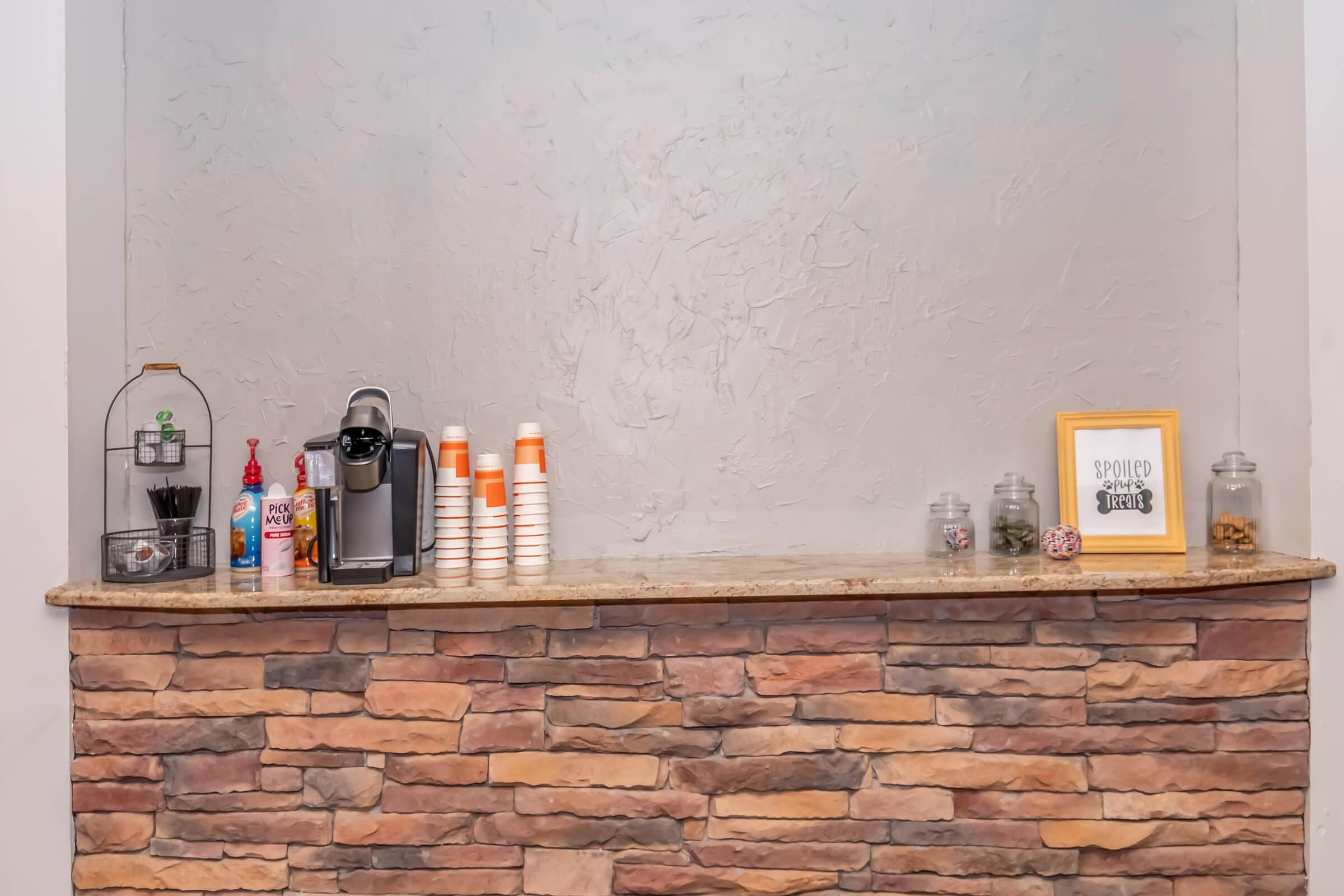 A countertop with a stone facade featuring a coffee machine, stacked cups, and various condiments. There are jars and a decorative frame with text on the right side. The wall behind is painted in a light gray tone, giving a clean and modern appearance.