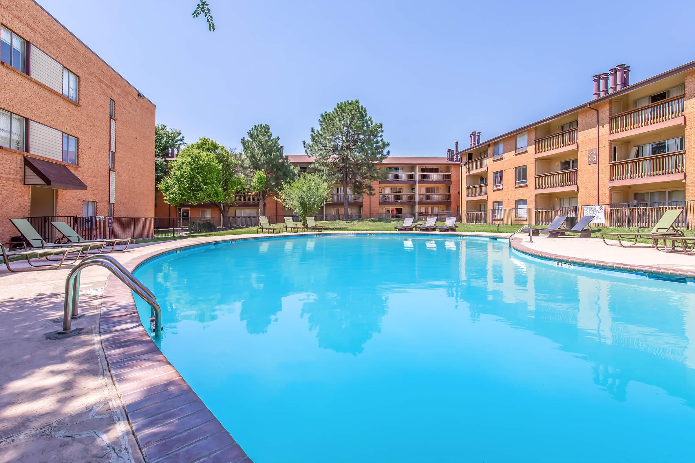 A spacious outdoor swimming pool in an apartment complex, surrounded by lounge chairs and leafy trees. The pool reflects a clear blue sky, and brick buildings with balconies are visible in the background, creating a relaxing atmosphere.