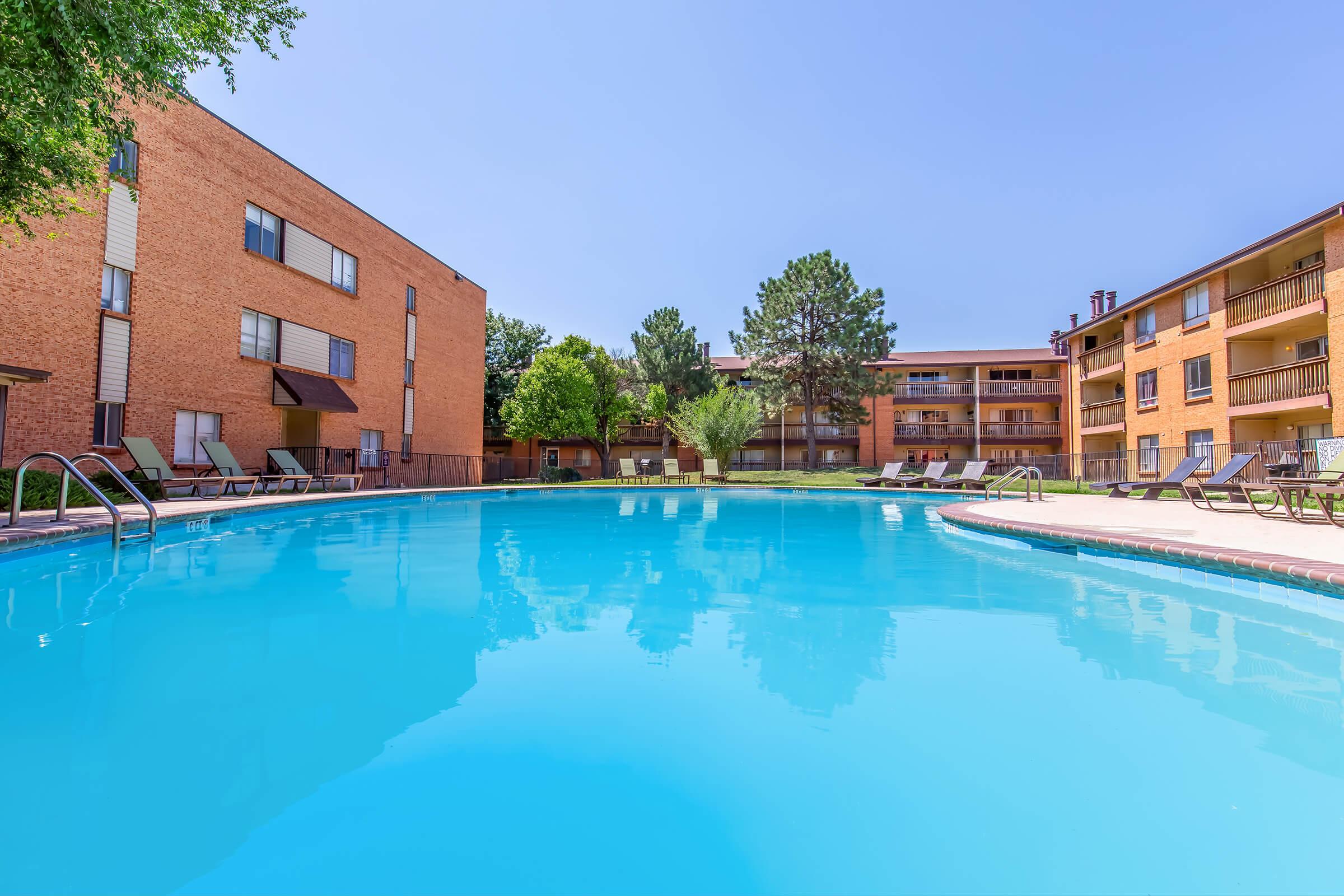 A clear blue swimming pool surrounded by lounge chairs, with landscaped greenery and buildings in the background. The scene is sunny, showcasing a peaceful outdoor space ideal for relaxation.