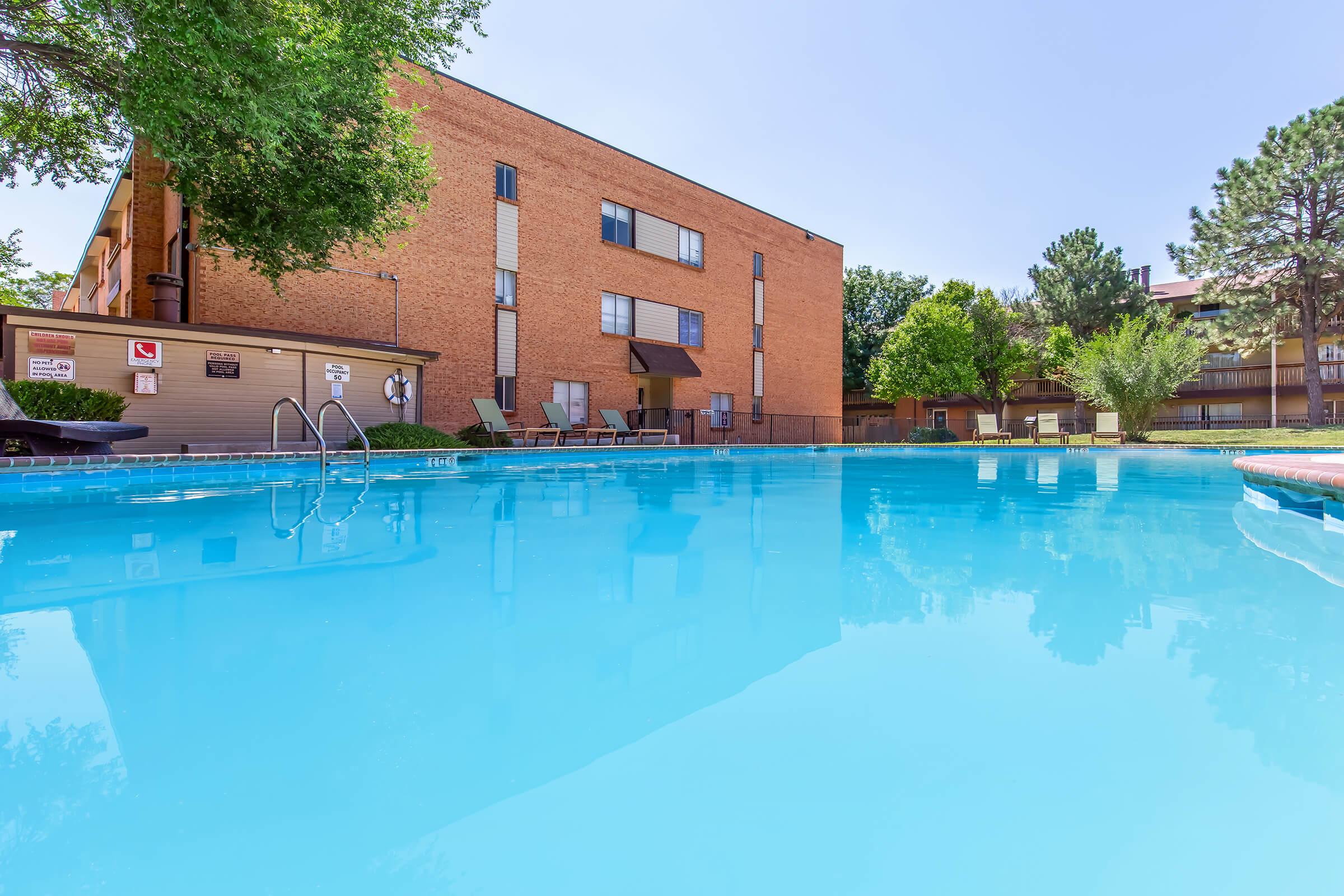 A clear blue swimming pool surrounded by lounge chairs, with a brick building and trees in the background. The sky is bright and sunny, reflecting off the water's surface.