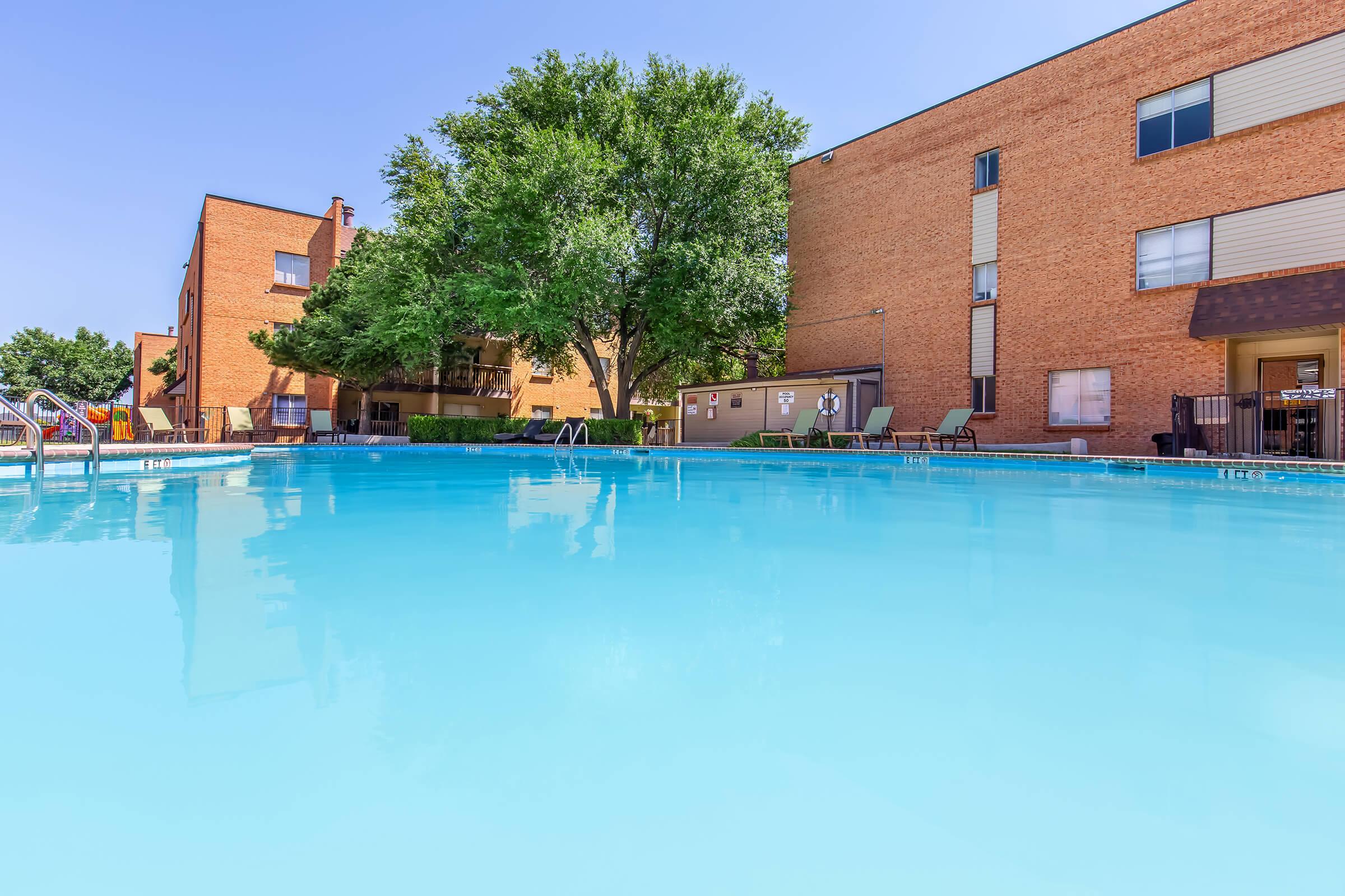 A clear blue swimming pool surrounded by green trees, with several lounge chairs and a brick apartment building in the background. The scene is bright and inviting, perfect for relaxation on a sunny day.