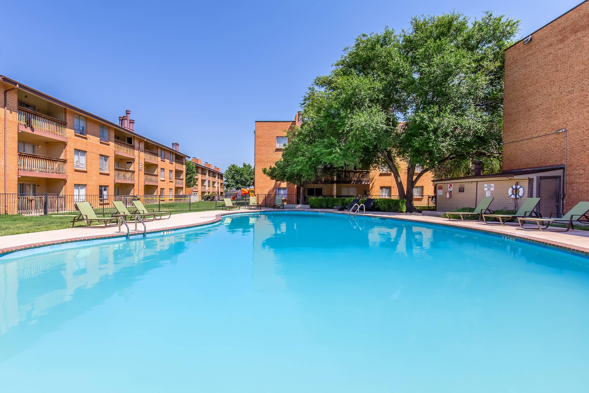 A bright outdoor swimming pool surrounded by lounge chairs and shaded by trees, with brick apartment buildings in the background under a clear blue sky. The pool area appears inviting and is well-maintained, suggesting a relaxing atmosphere.