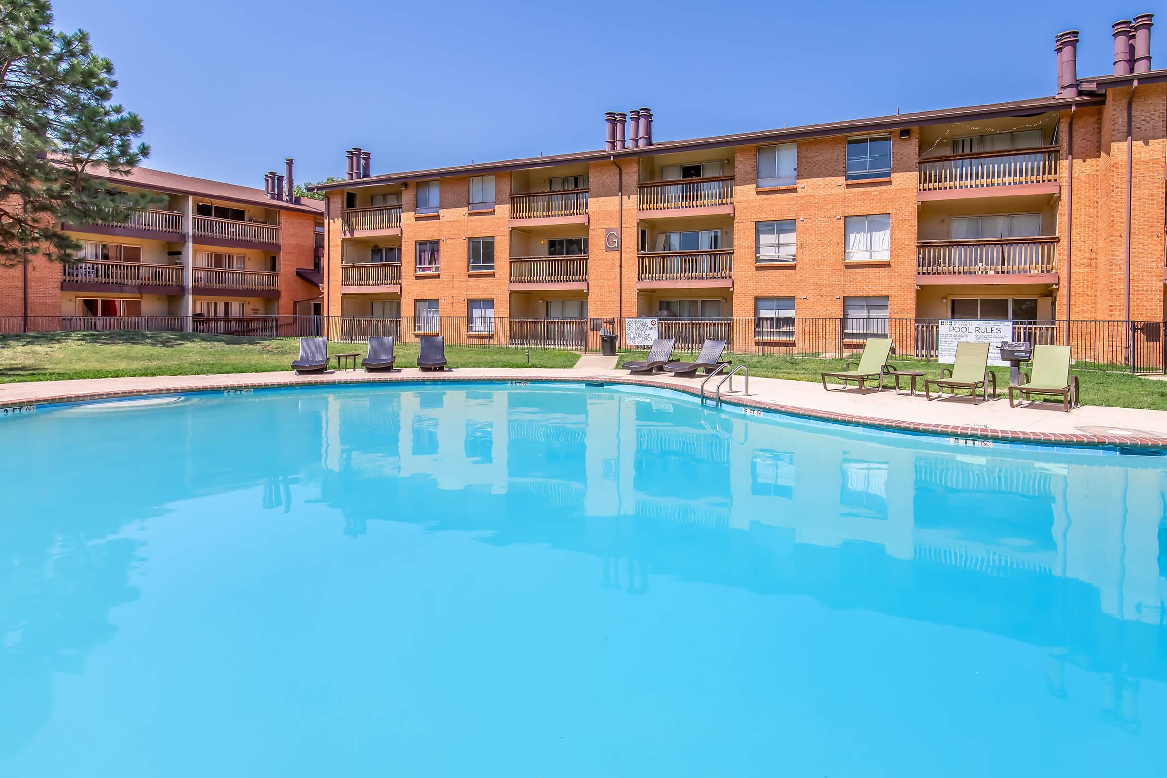 A clear blue swimming pool reflects the surrounding orange brick apartment buildings on a sunny day. Lounge chairs line the poolside, and green grass is visible in the background, creating a relaxing outdoor atmosphere.