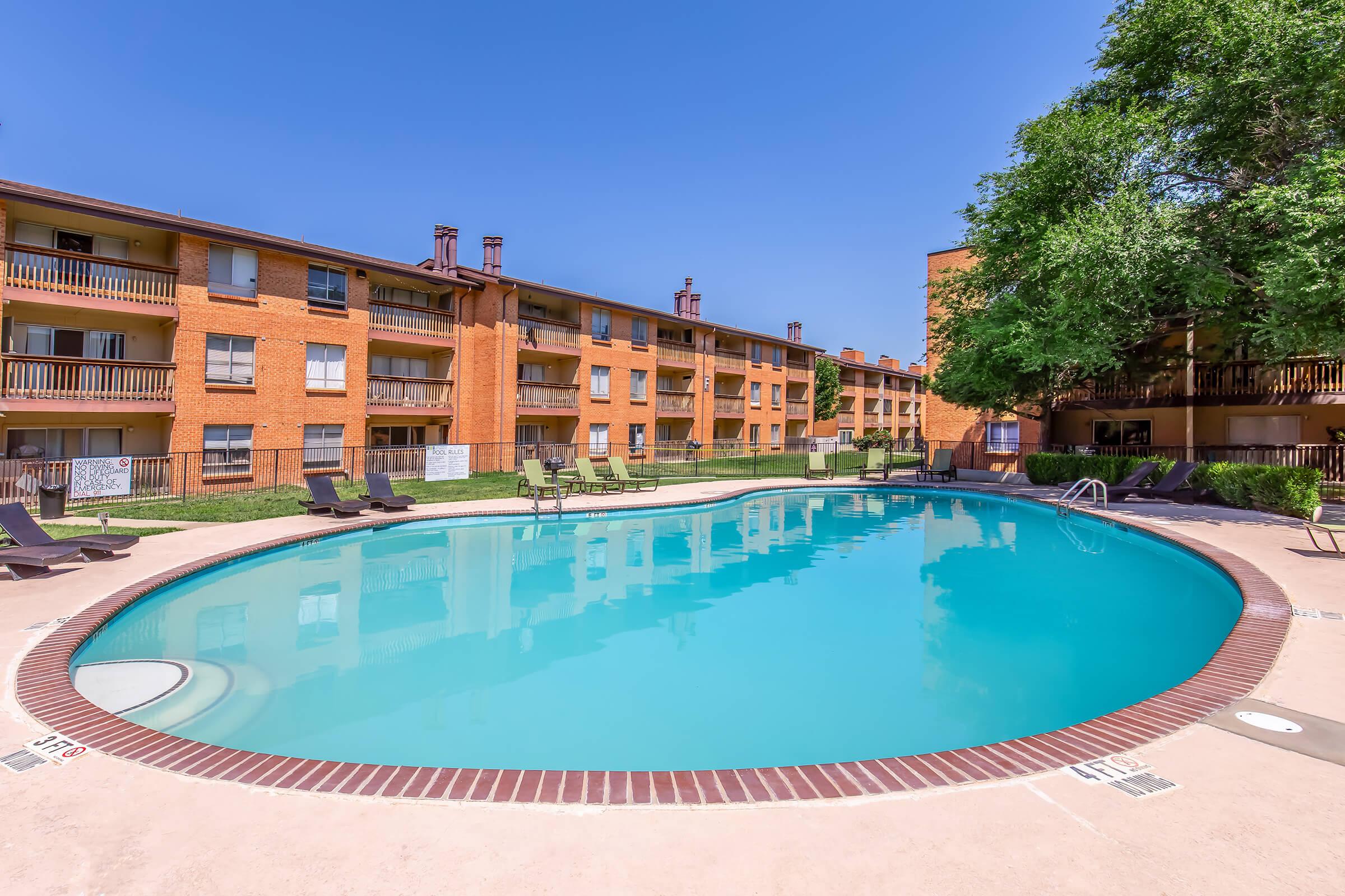 A clear swimming pool surrounded by lounge chairs, with two orange brick apartment buildings in the background. The scene features a bright blue sky and well-maintained greenery around the pool area, creating a relaxing outdoor atmosphere.