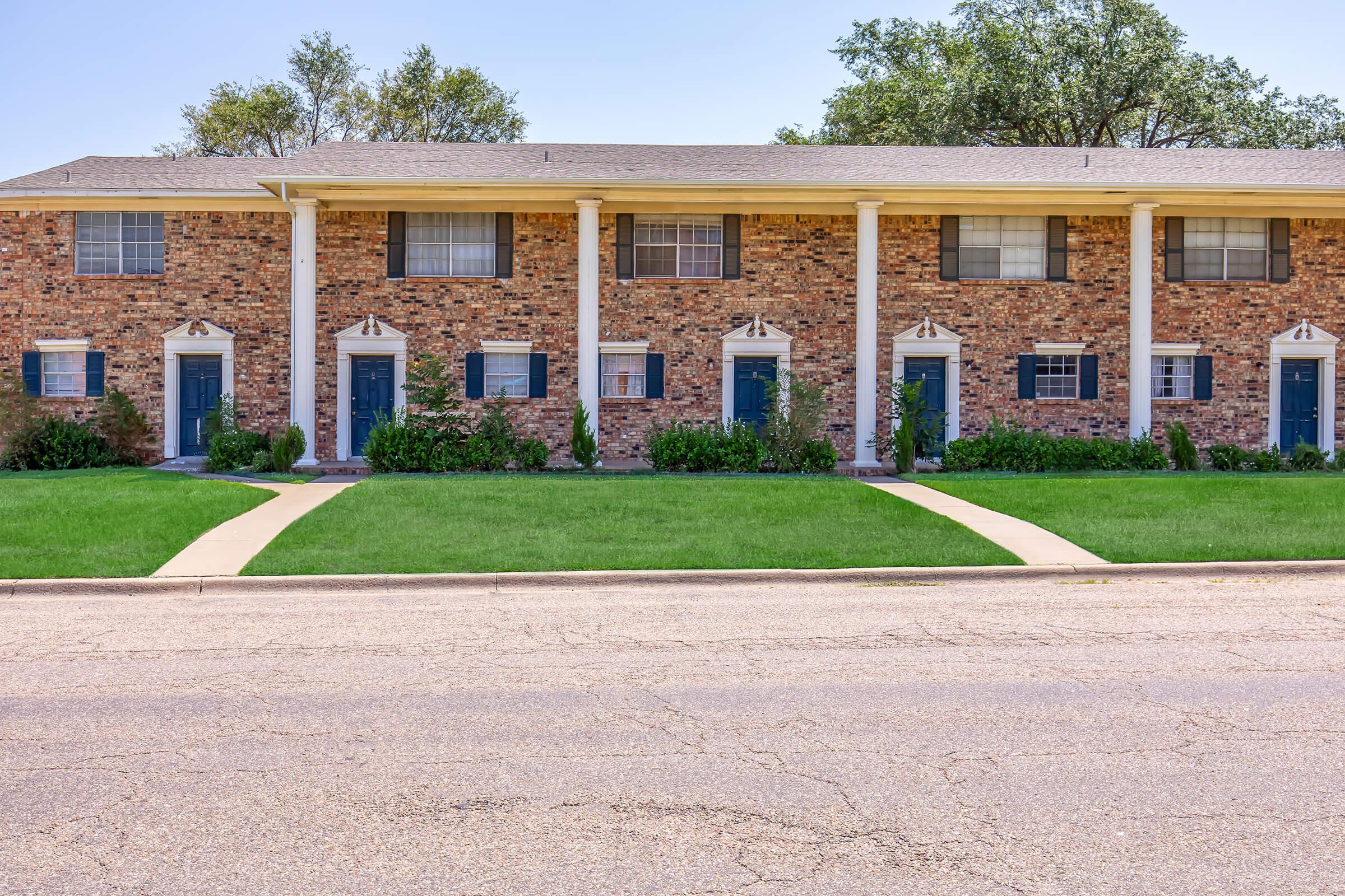 Row of brick townhouses with blue doors and white columns, surrounded by well-maintained lawns and shrubs. Sidewalks lead up to the entrances, and the scene is set under a clear blue sky, portraying a pleasant residential environment.