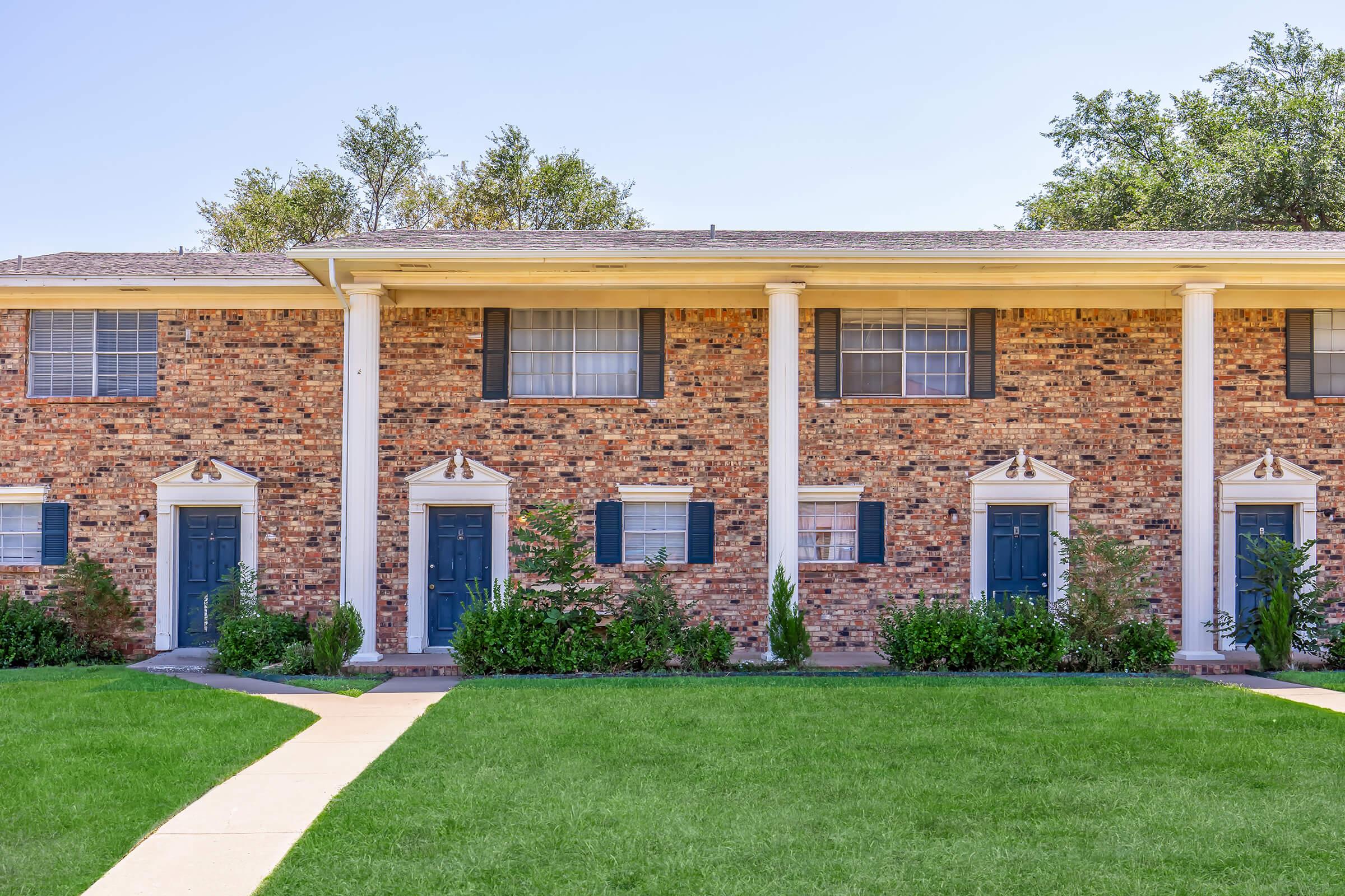 A brick apartment building with three identical units, each featuring blue doors and white trim. There are decorative pillars by the entrances and well-maintained greenery in front. A paved walkway leads to the doors, set against a clear blue sky.