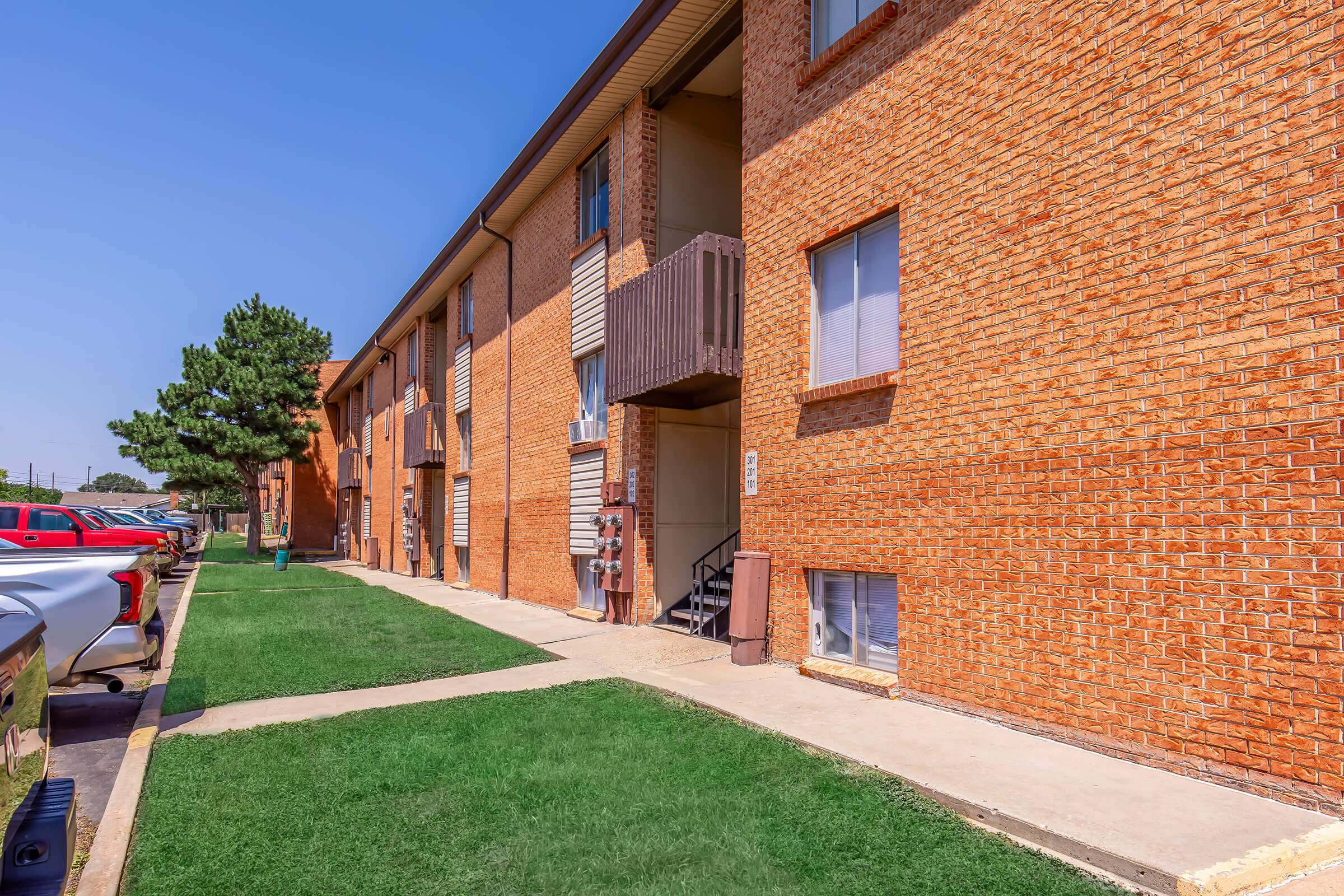 Exterior view of a brick apartment building with several windows and a staircase. The ground is grassy, and nearby, there are parked vehicles. The setting is clear and sunny, highlighting the architecture of the building and the surrounding area.