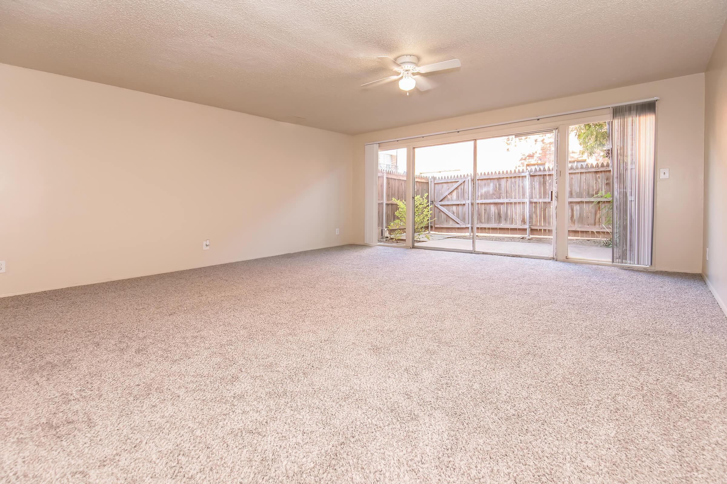 A spacious indoor room with light beige walls and a large, empty carpeted area. A ceiling fan is visible, and a sliding glass door leads to a patio area with a wooden fence and greenery outside. The room is bright and uncluttered, suggesting a fresh, open living space.