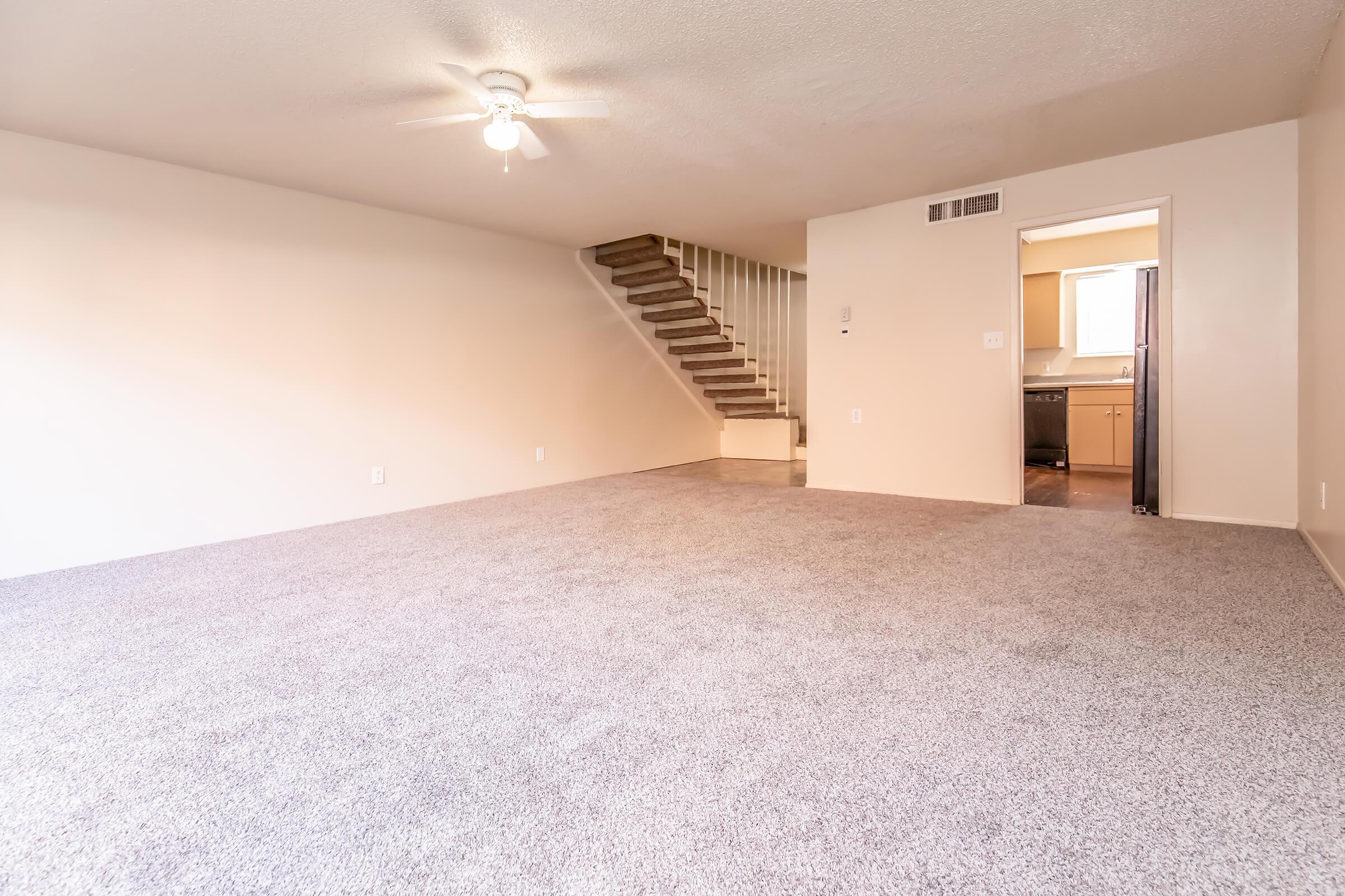 Spacious living area with light gray carpet, featuring a ceiling fan. Stairs lead to a second floor, and a doorway in the background opens to a kitchen with modern appliances. The walls are painted a soft beige, enhancing the bright and airy atmosphere of the room.