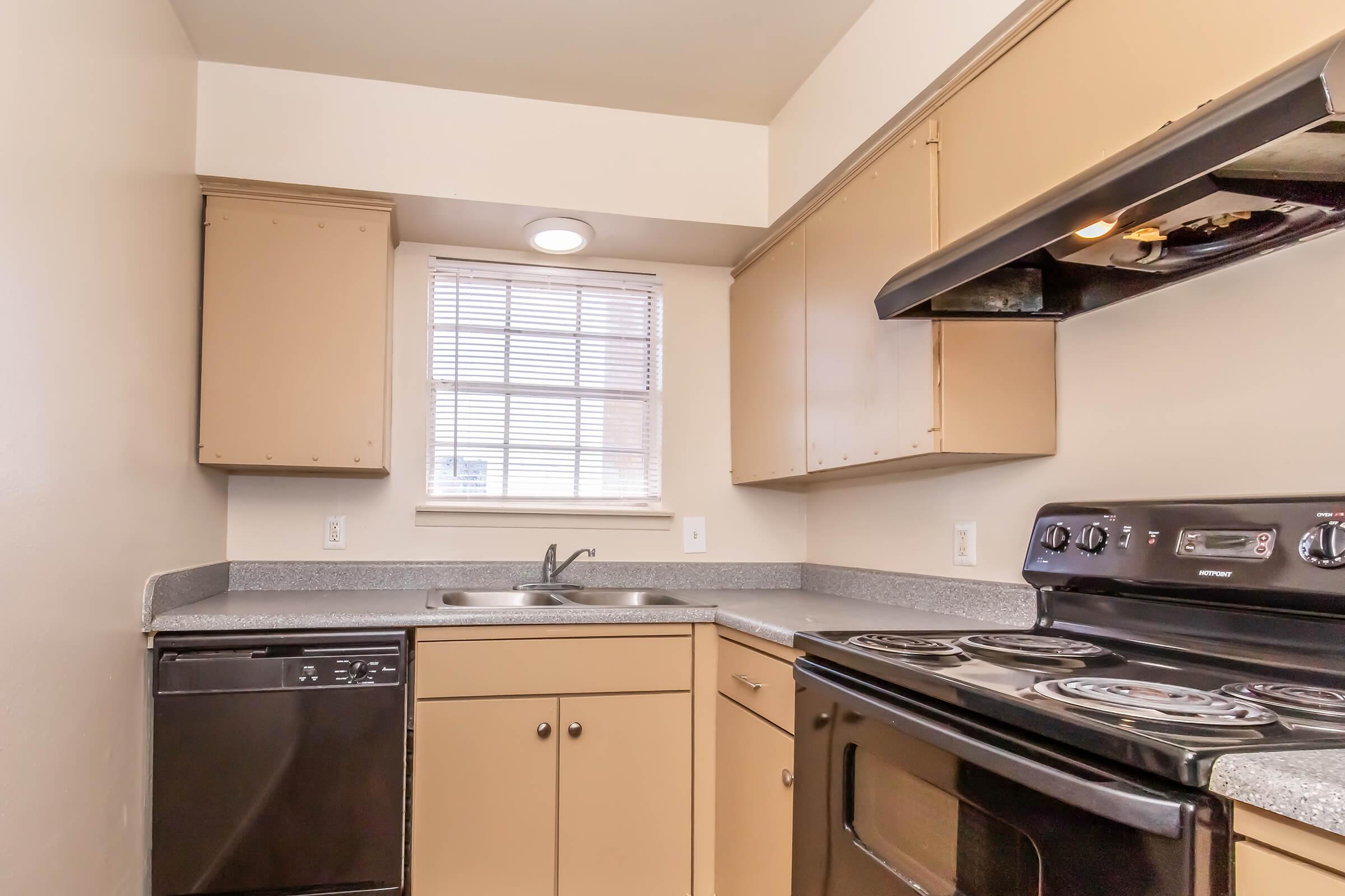 A kitchen with beige cabinets, a black stove, and a dishwasher. The countertop is gray, and a window provides natural light. The layout is simple, with the sink located near the window and appliances neatly arranged.