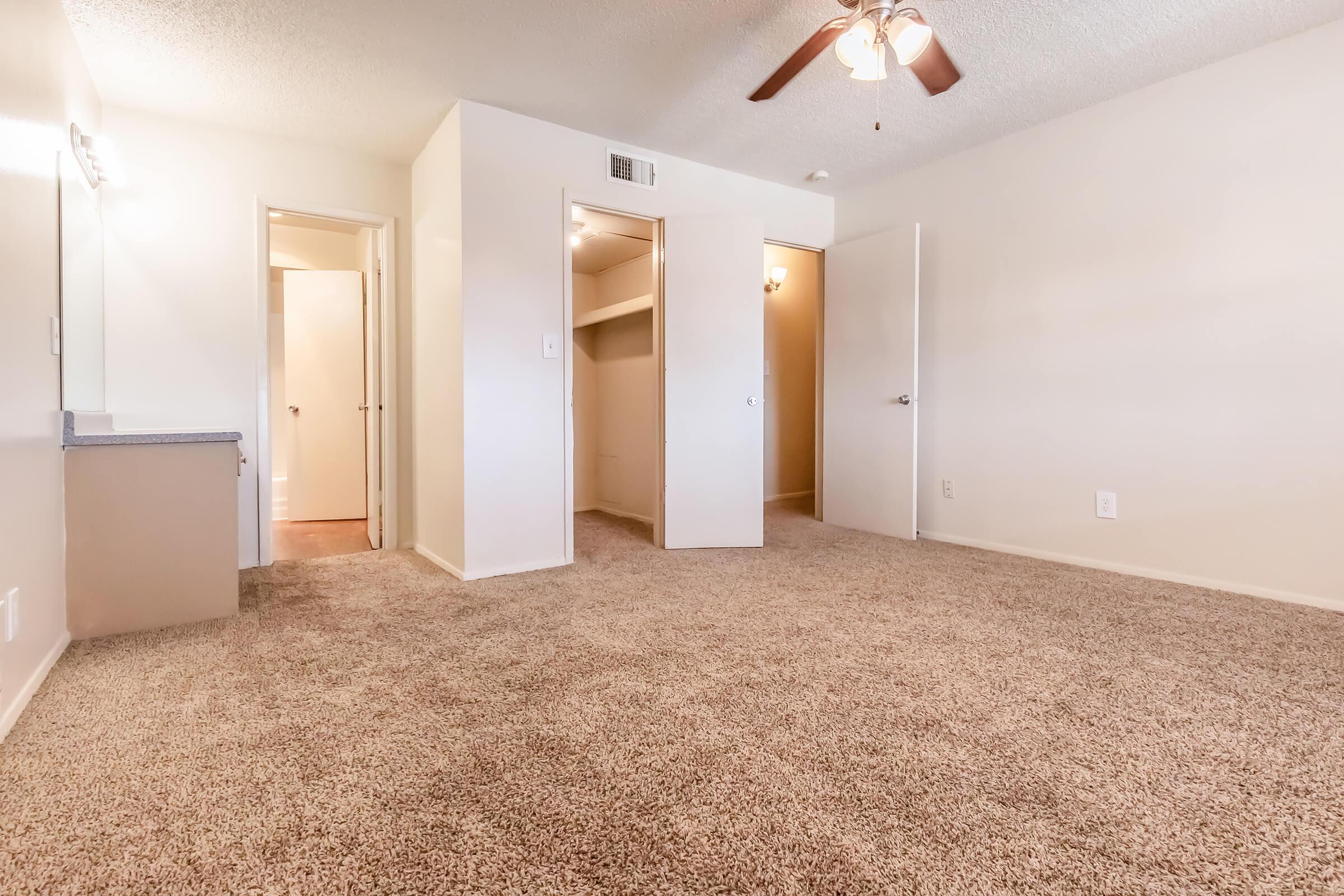 A well-lit, empty room featuring carpeted flooring, a ceiling fan, and multiple doorways leading to closets and other spaces. The walls are painted in a neutral tone, creating a spacious and inviting atmosphere.
