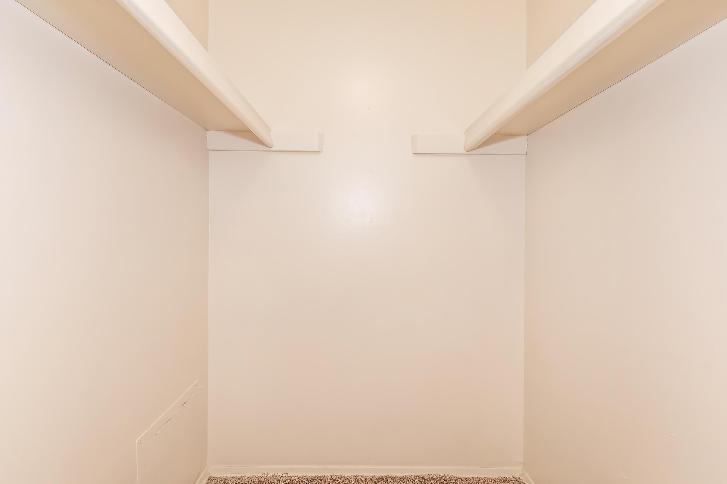 Empty closet with light beige walls and two horizontal shelves. The floor is covered with a light-colored carpet, and there are no items or decorations present, creating a minimalist look.