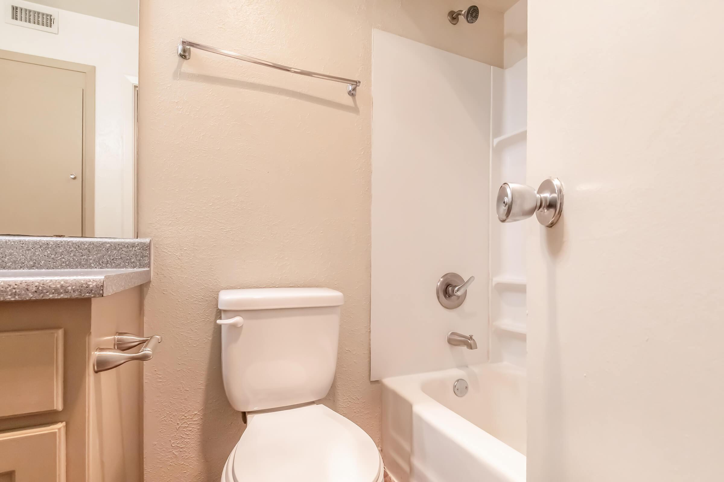 A clean, modern bathroom featuring a white toilet, bathtub with shower fixture, and a simple vanity with a countertop. The walls are painted in a light color, and there's a towel bar mounted on the wall. The overall aesthetic is minimalist and functional.