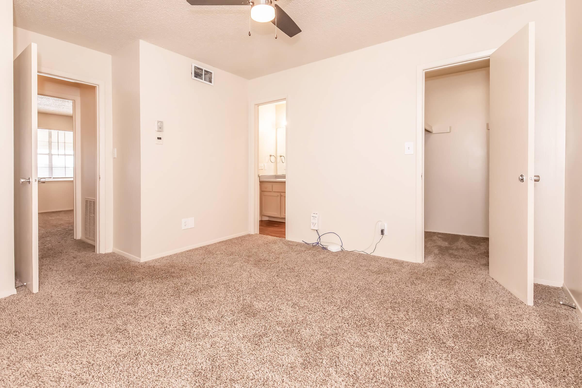 A spacious, neutral-colored room with carpet flooring, featuring two open doorways leading to adjacent areas. There is a ceiling fan above, and the light-colored walls create a bright atmosphere. The doorway on the right leads to a closet, while the left opens to another room.
