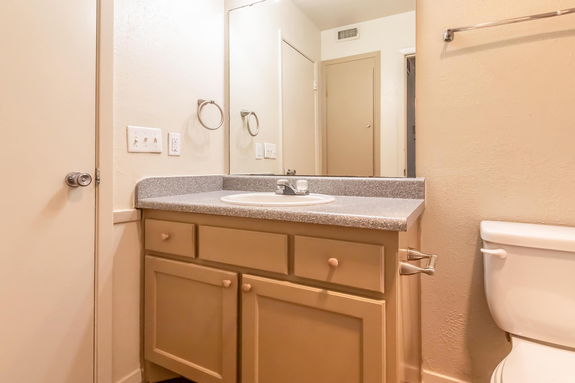 A bathroom featuring a beige countertop with a sink, a mirror, and a towel ring. The cabinetry below the sink is also beige. A white toilet is positioned to the right, and there is a door leading to another area in the background. The walls are painted a light color, giving the space a clean and simple appearance.