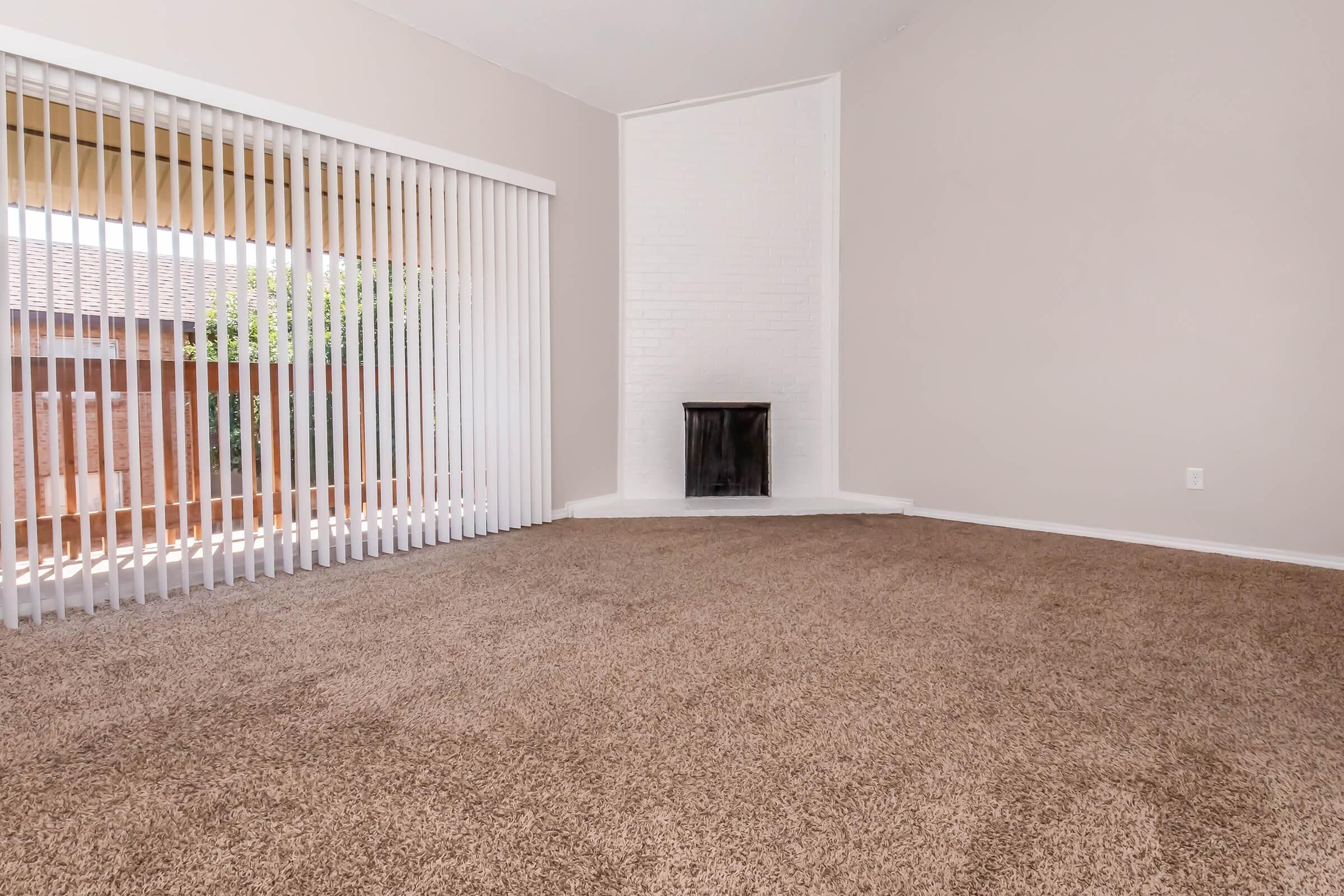 Spacious, empty room with beige walls and light brown carpet. There is a white brick fireplace in the corner and large sliding glass doors with vertical blinds allowing natural light. The overall atmosphere is bright and inviting, perfect for a cozy living area or family room.