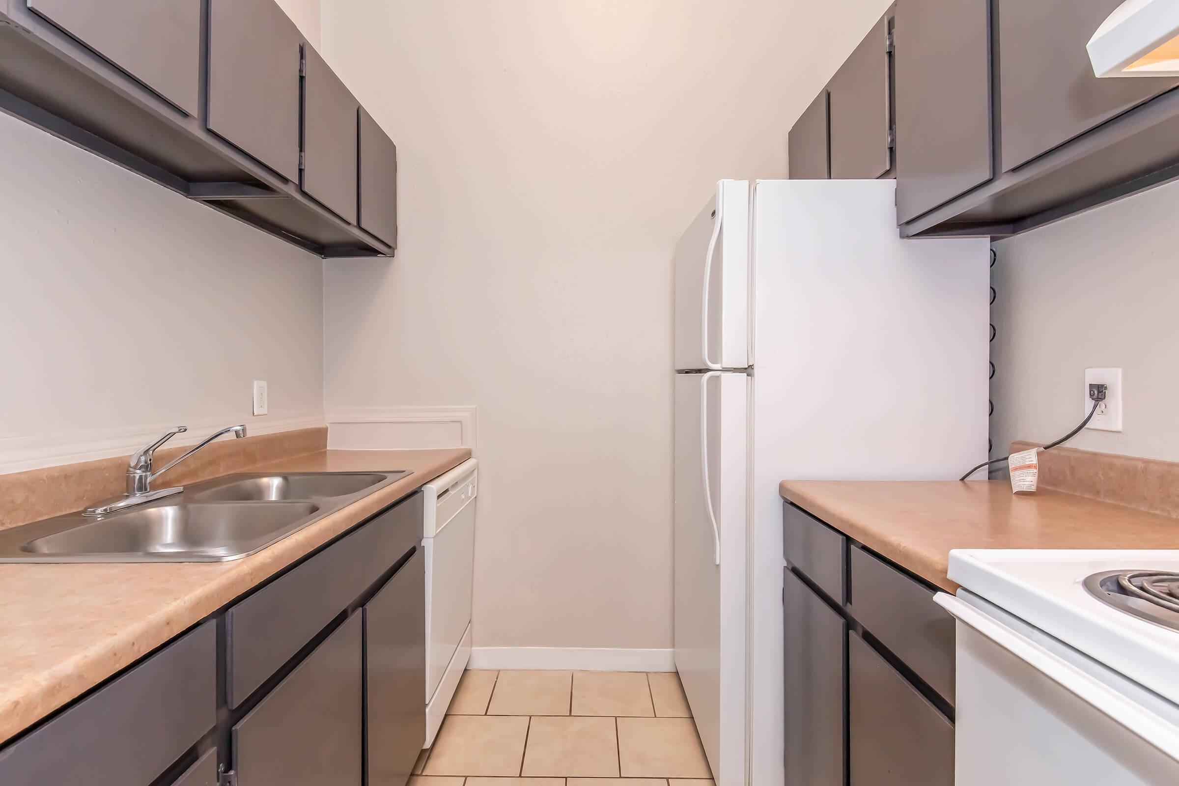 A modern kitchen featuring dark cabinets, a double sink, a dishwasher, a white refrigerator, and a stove. The countertops are a light beige, and the floor is tiled in a similar shade. The walls are painted in a soft neutral color, giving the space a clean and inviting look.