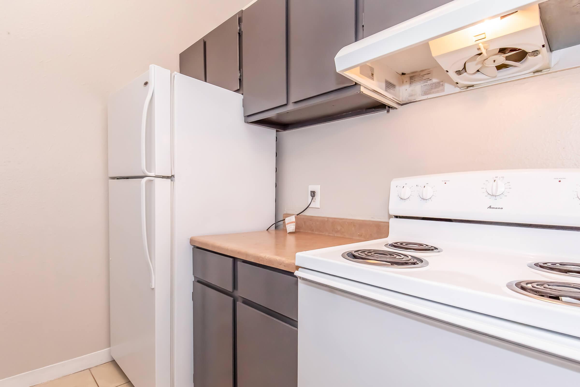 A kitchen corner featuring a white electric stove with an oven, a white refrigerator, and dark wood cabinetry. The countertops are beige, and the walls are light-colored. The setup is clean and functional, ideal for cooking and food preparation.