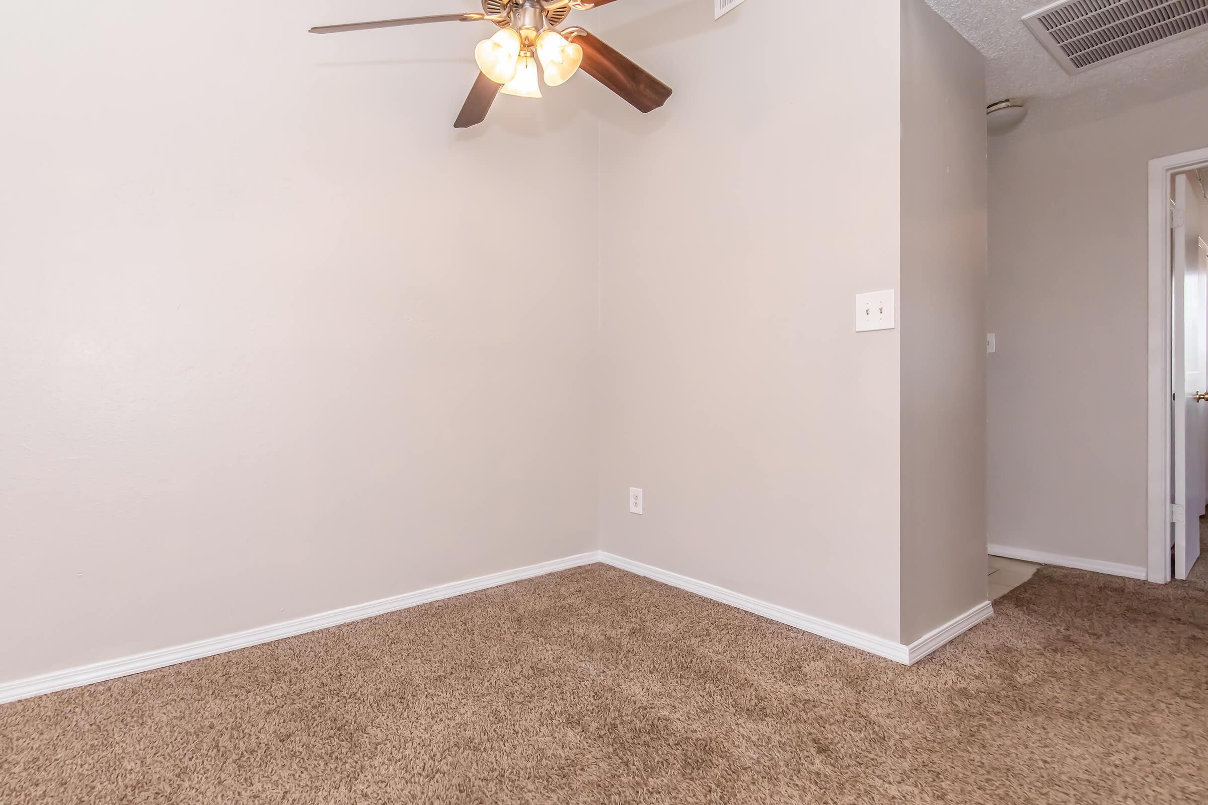 Empty interior room featuring light beige walls and a ceiling fan with four bulbs. The floor is covered in tan carpet, and there is a doorway visible in the background. The space appears well-lit and uncluttered, suitable for various uses.