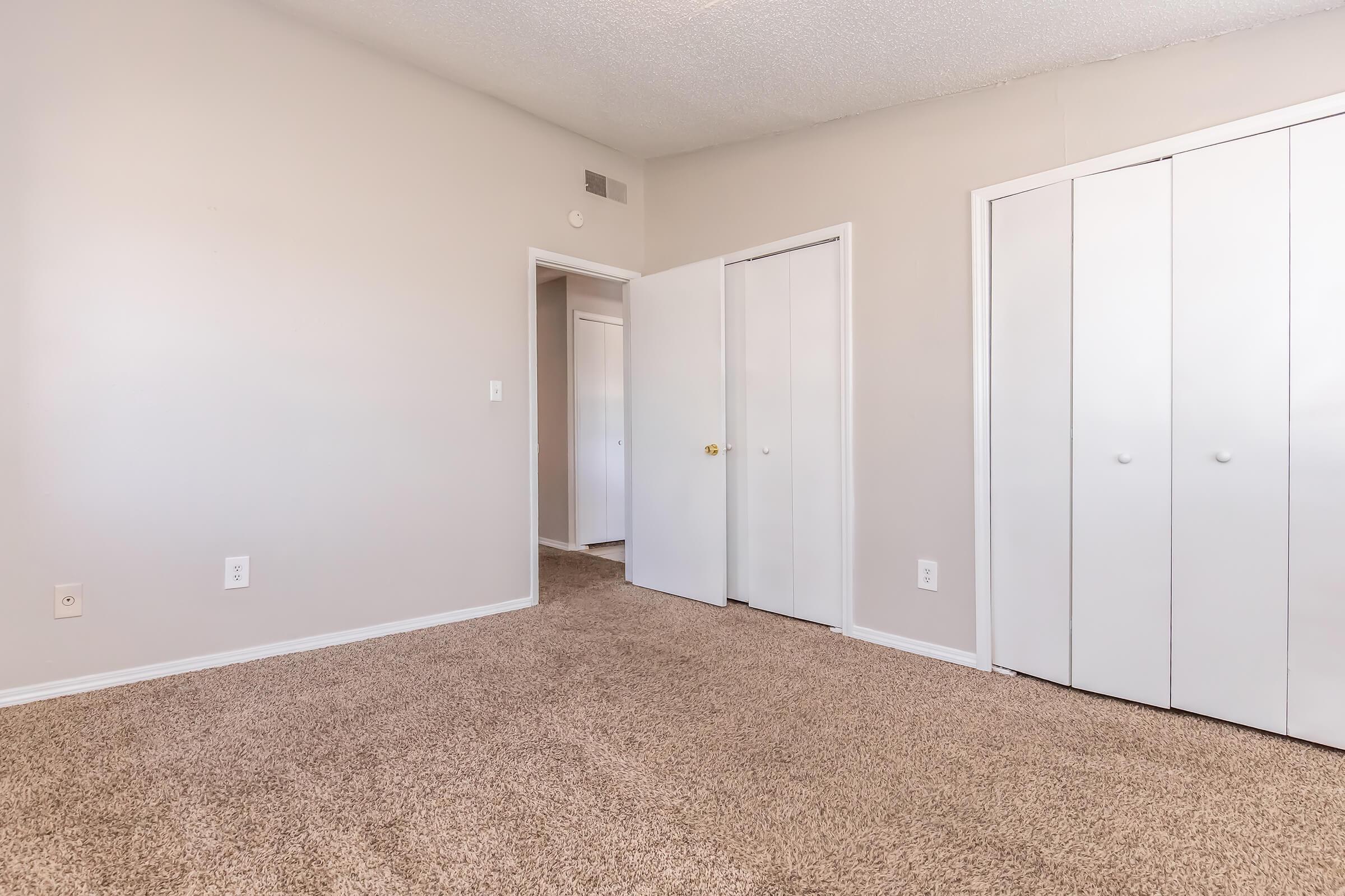 A well-lit, empty room with beige carpet and light gray walls. There are two doors, one leading to a closet with white sliding doors and another to a hallway. The space appears clean and tidy, suitable for various uses such as a bedroom or an office.