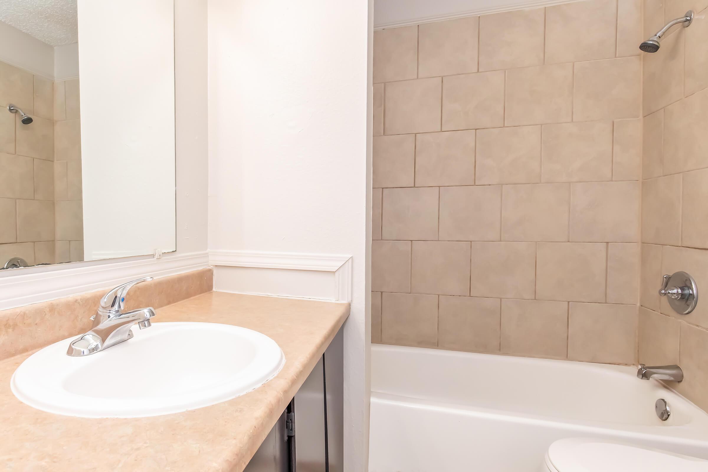 A bathroom featuring a vanity with a sink and mirror, beige tile walls, and a bathtub with a shower. The design is simple and clean, emphasizing functionality and neutral tones.