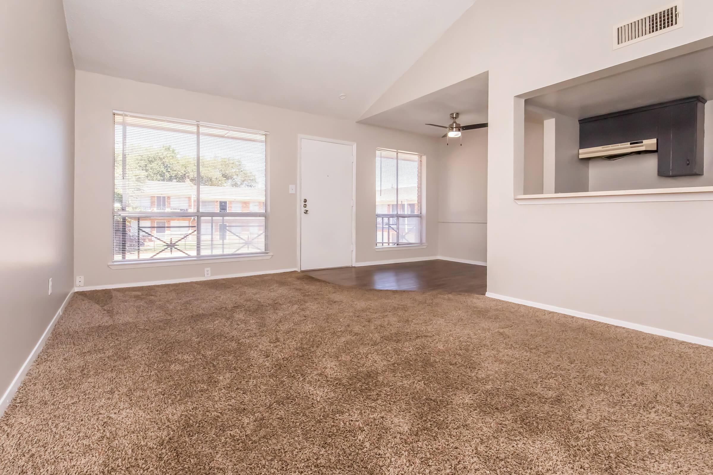 A spacious and airy living room with light gray walls and plush brown carpet. Large windows allow natural light to fill the room, and a ceiling fan hangs from the vaulted ceiling. An open layout leads to a partial wall that features an opening to a kitchen area.
