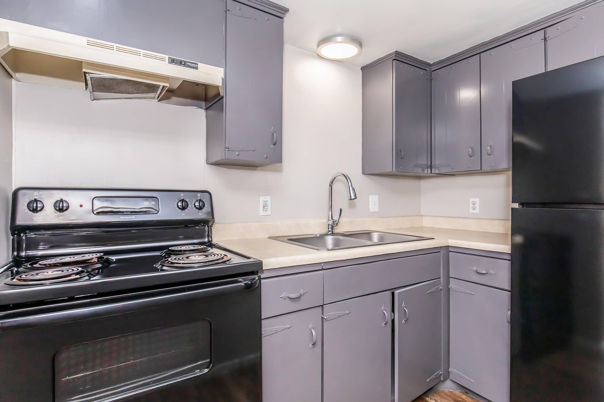 A modern kitchen featuring dark gray cabinets, a black stove with a hood above, a stainless steel sink, and a black refrigerator. The countertop is light-colored, and there is overhead lighting, creating a clean and functional cooking space.